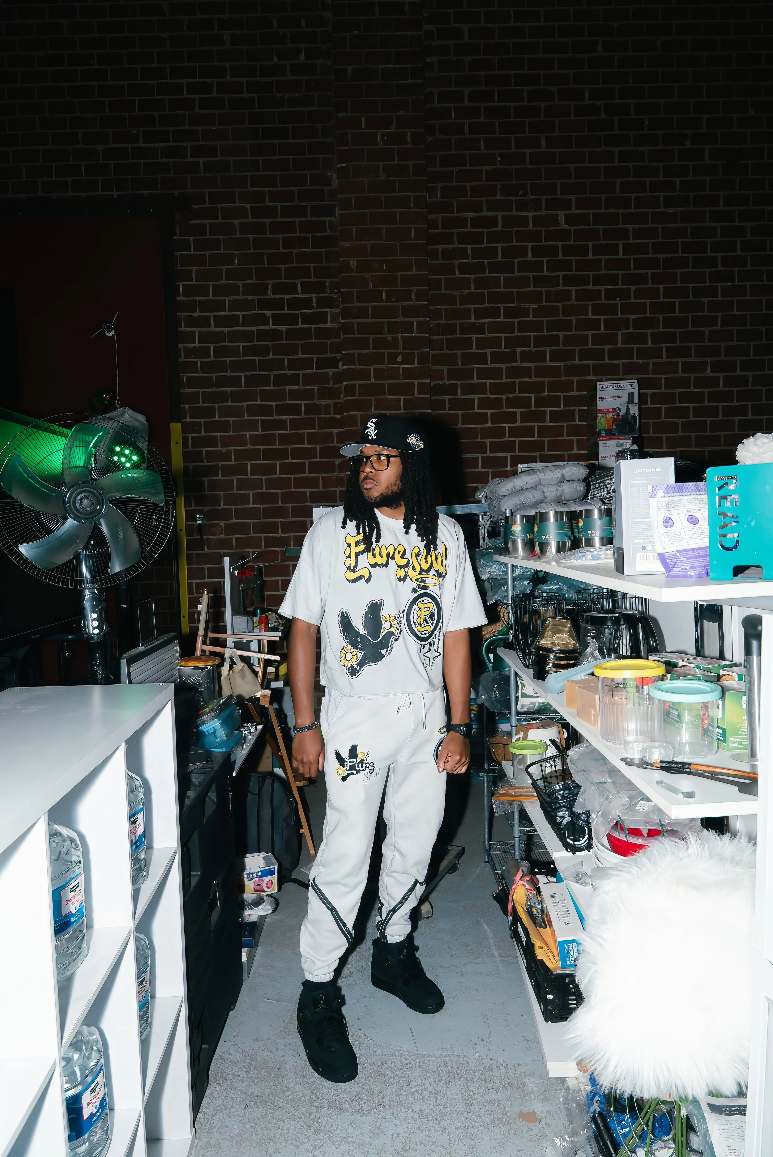 A man with glasses and dreadlocks wearing a white graphic t-shirt and sweatpants stands in a storage or supply room surrounded by shelves filled with kitchenware, containers, and miscellaneous items.