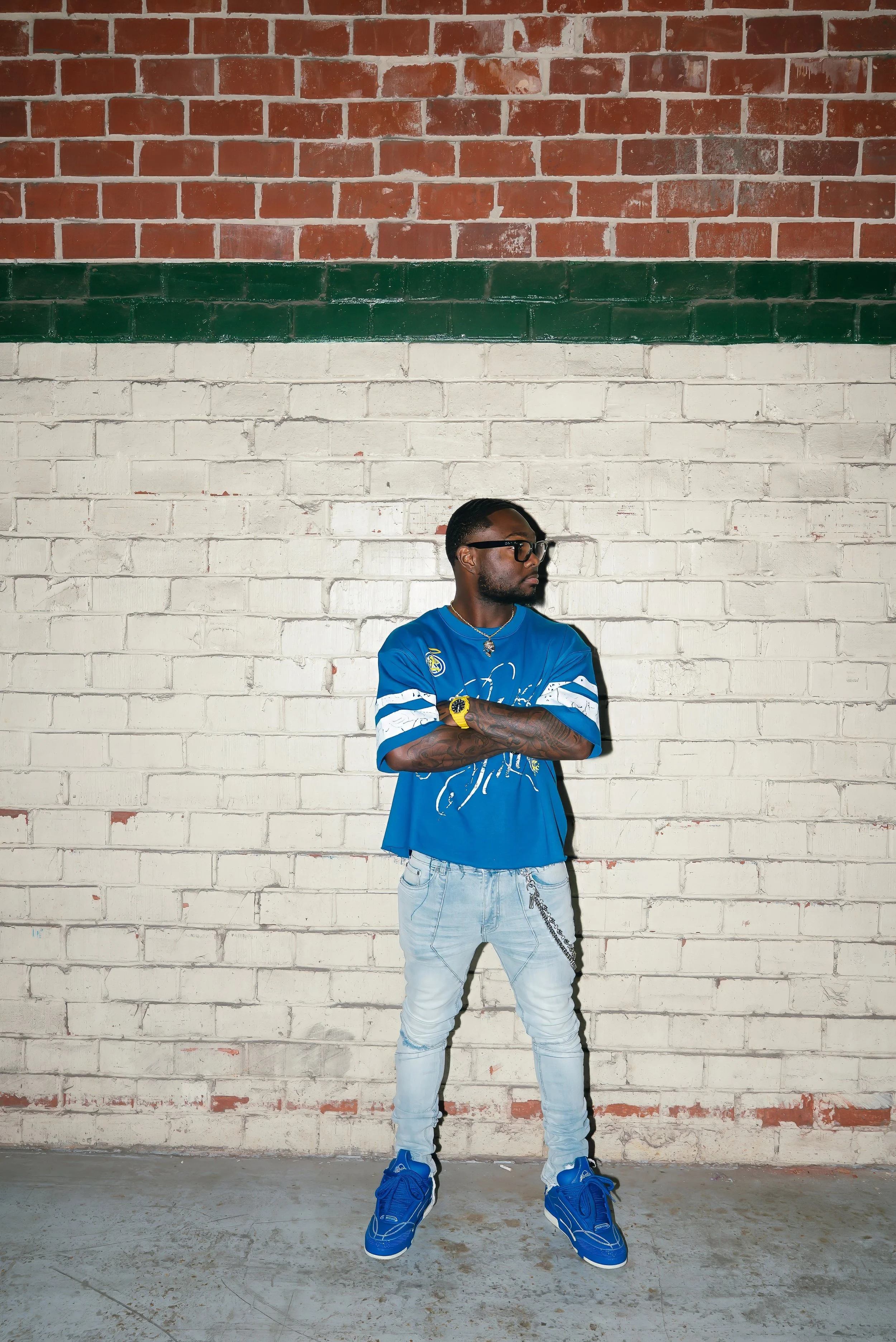 A man standing with arms crossed in front of a brick wall painted with horizontal stripes in red, green, and white.