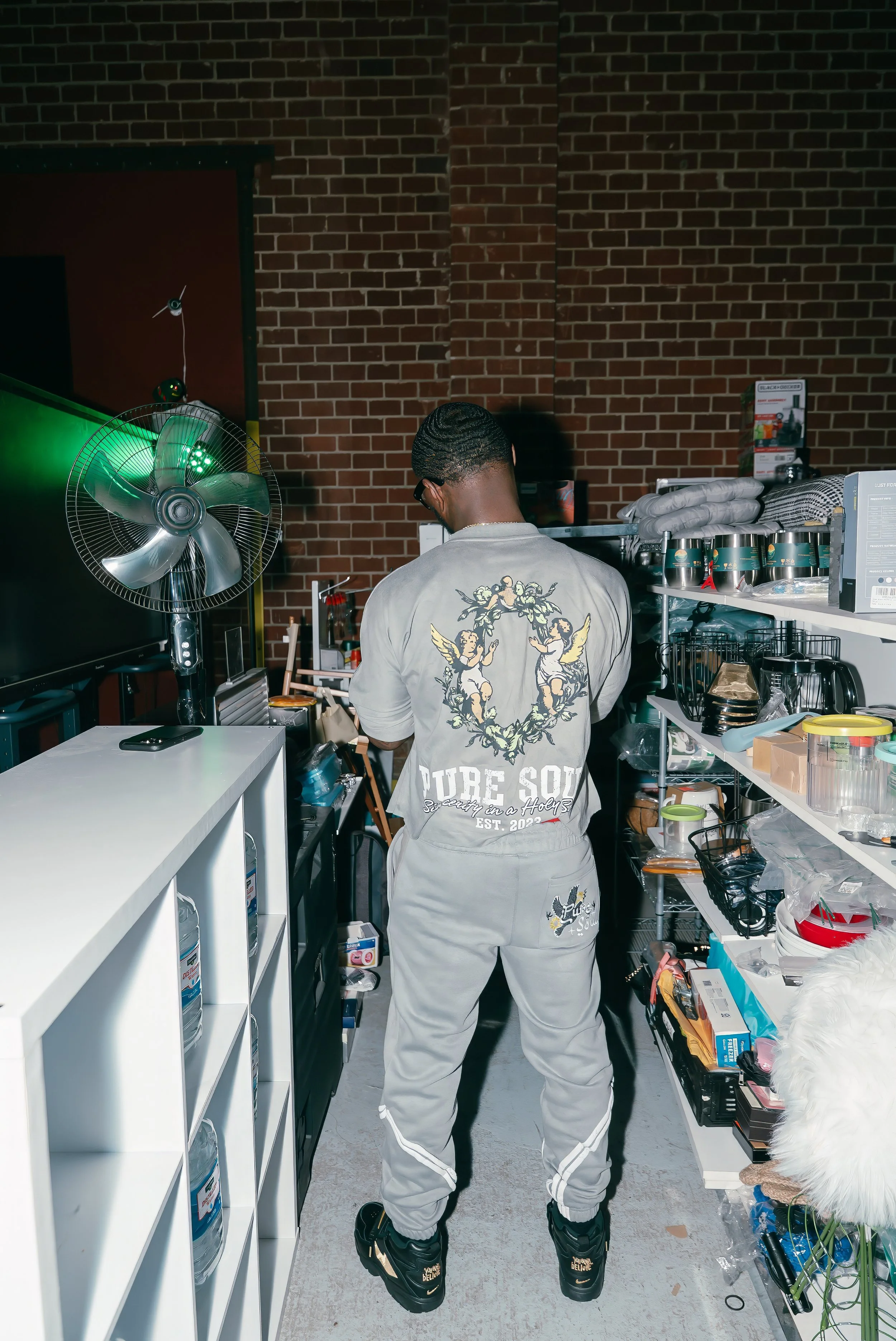 A person with short hair and glasses, wearing a matching light gray tracksuit with 'PURE SOUL' written on the back, standing in a store or storage room surrounded by shelves with various items, including cups, boxes, and miscellaneous objects.