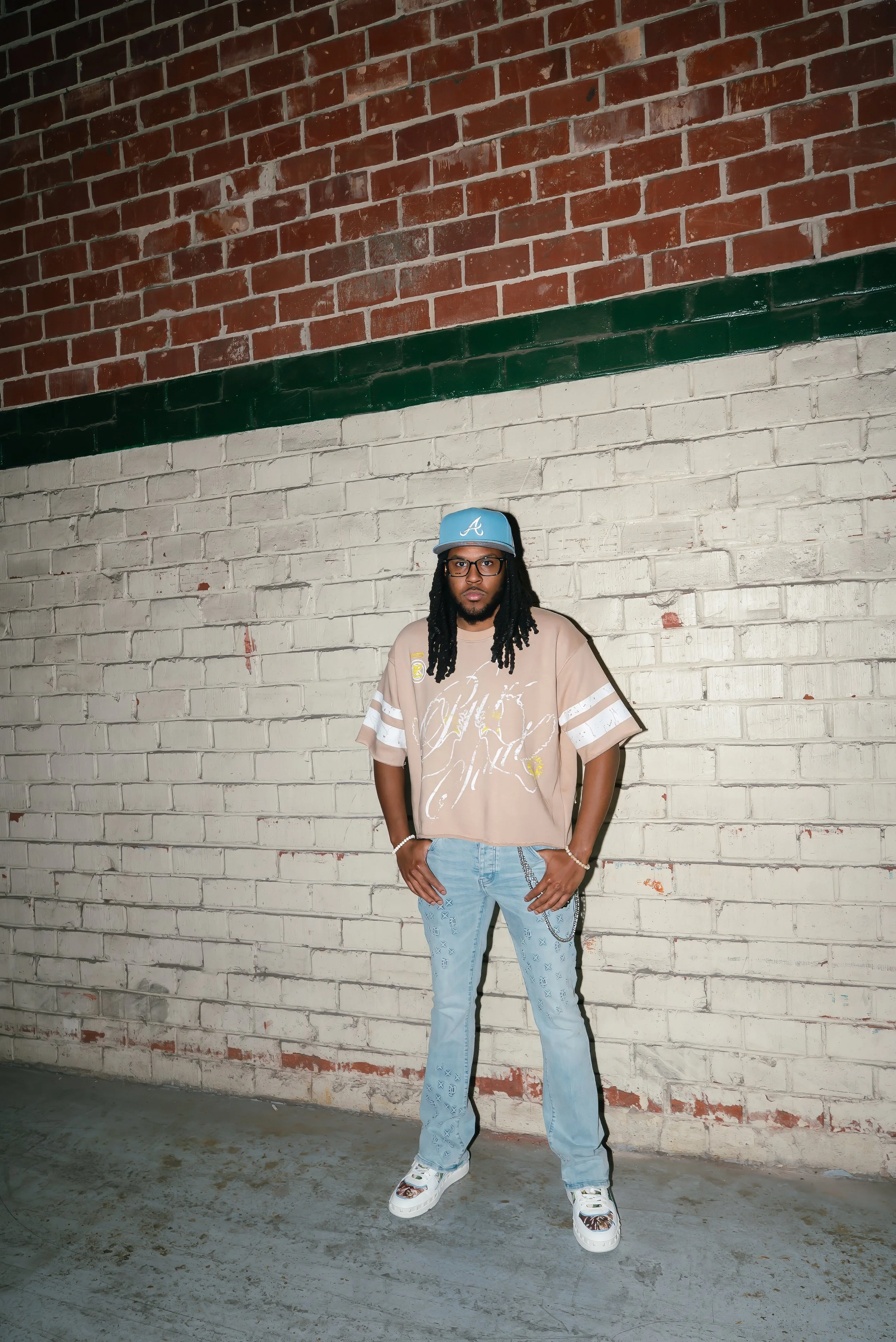 A young man with glasses, dressed in a beige graphic t-shirt, light blue jeans with embroidery, white sneakers, and a blue cap, stands against a brick wall with red, white, and green sections.