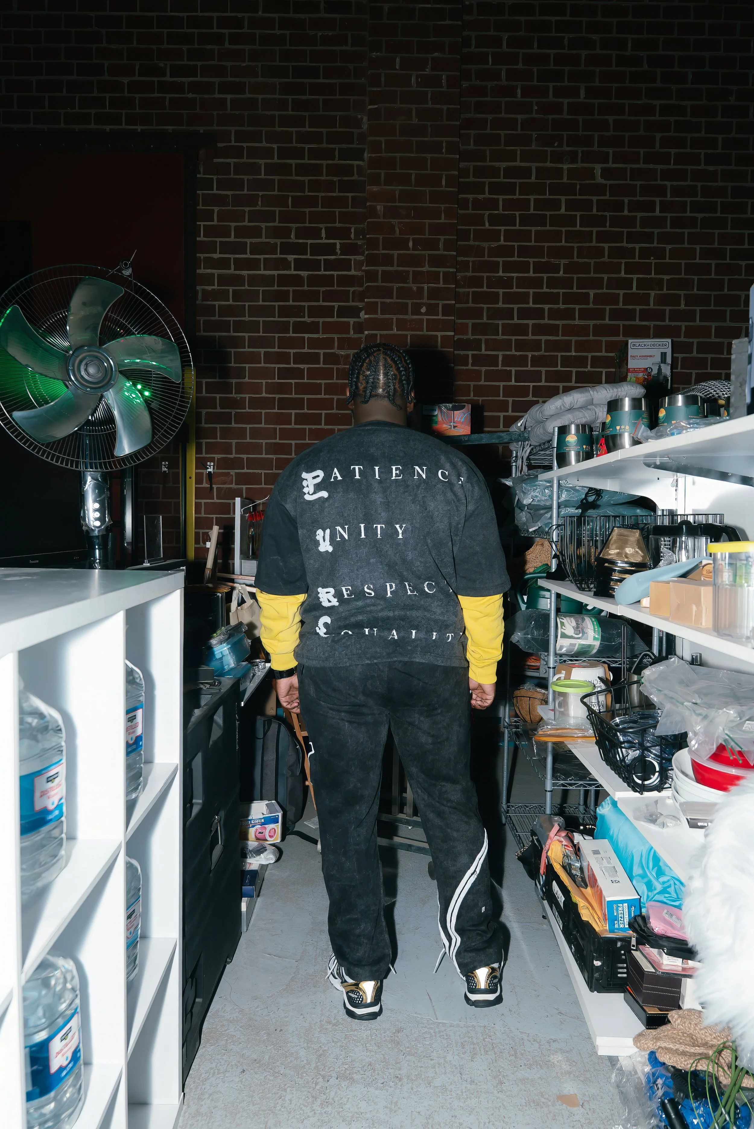 A person with braided hair is standing in a cluttered storage room or kitchen, facing away from the camera. They are wearing a black over-sized shirt with words on the back, yellow long-sleeve shirt underneath, black pants, and sneakers. The room has