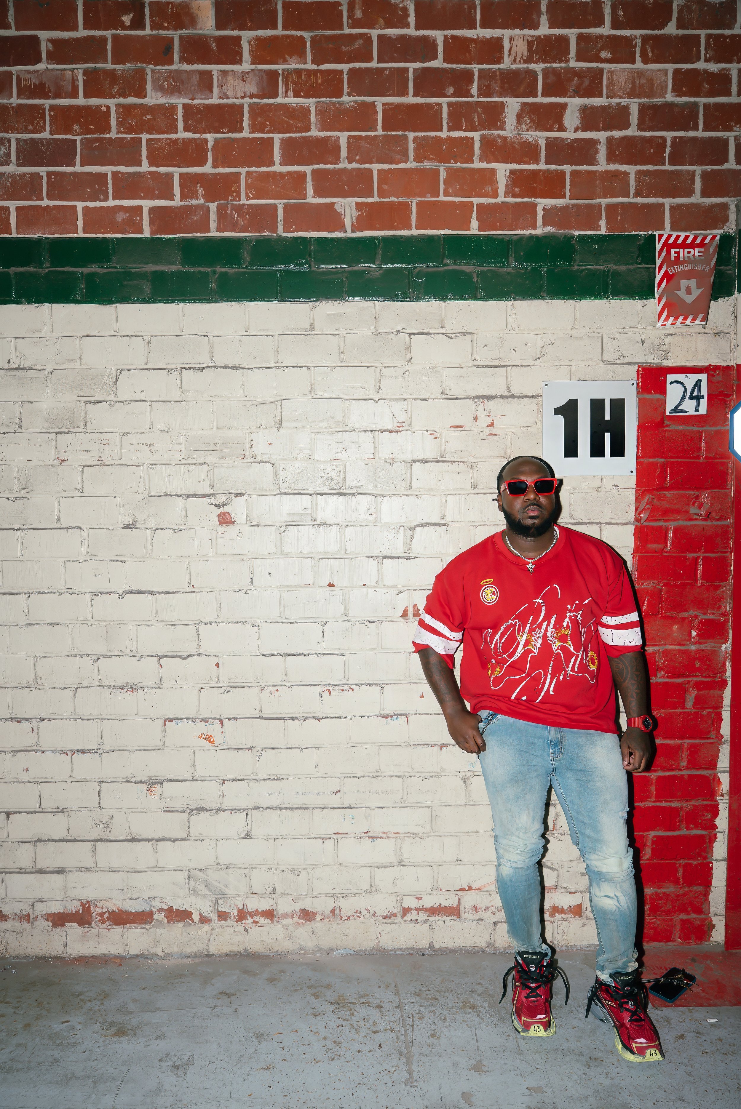 A man dressed in a red soccer jersey, light blue jeans, and red sneakers, wearing red sunglasses, standing in front of a brick wall with painted sections in red, white, and green, and signage including a fire extinguisher sign, a number "1H", and a n