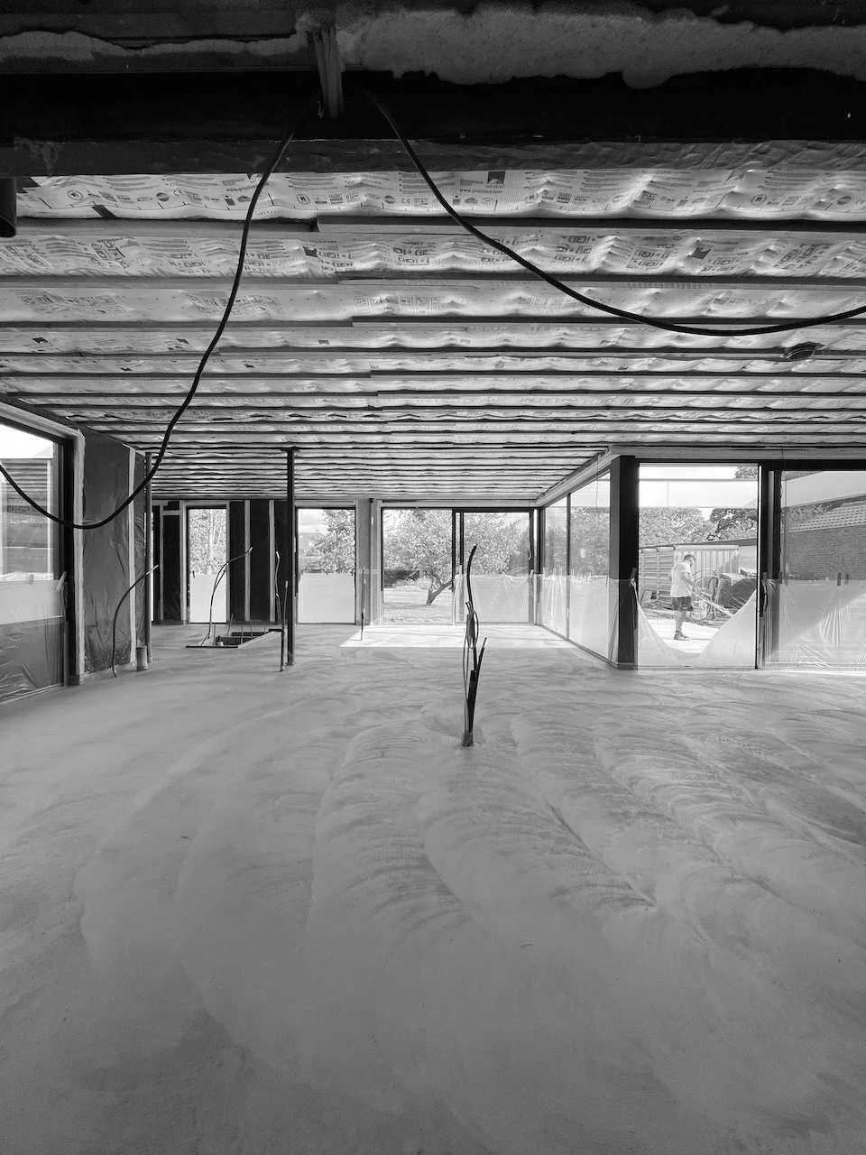 Binnenkant van een huis onder constructie met onafgewerkte vloer, muren en plafonds in aanbouw, en grote ramen naar buiten, waar mensen werken.