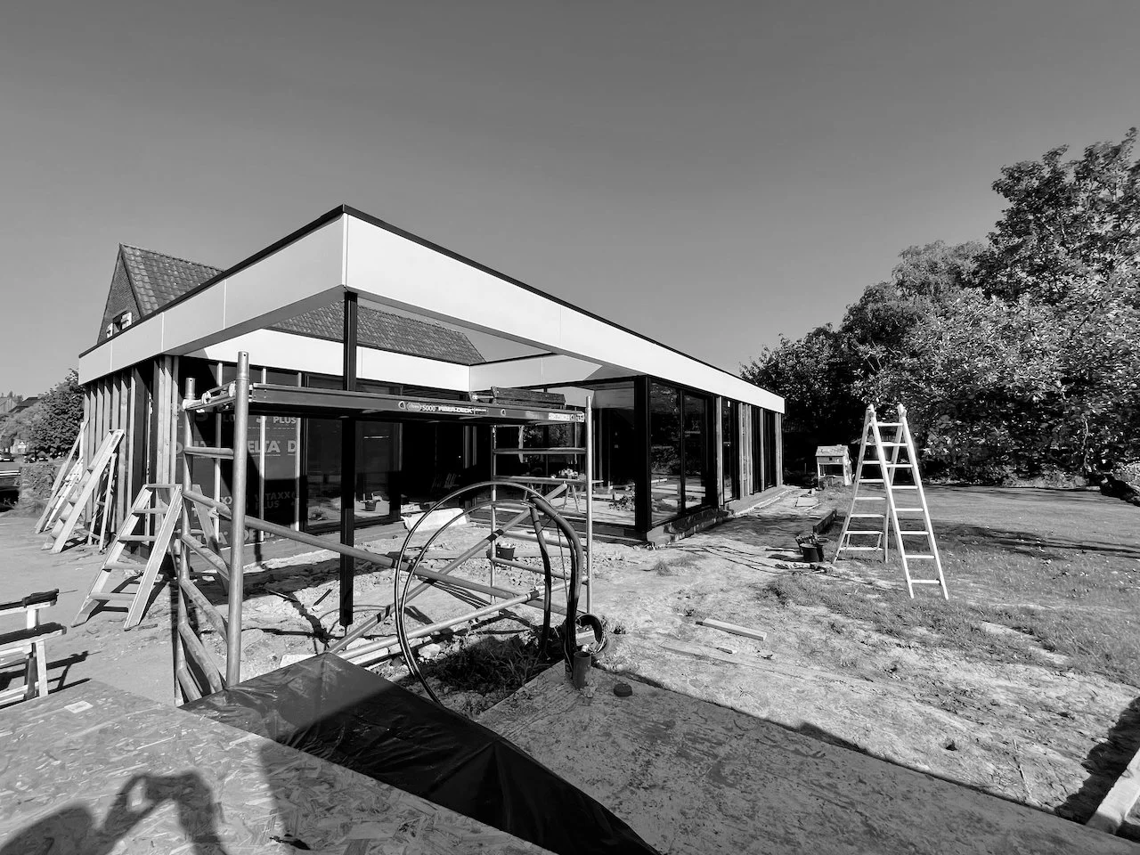 Moderne bouw van een huis onder constructie met steigers en ladders, omringd door bomen en een open terrein.