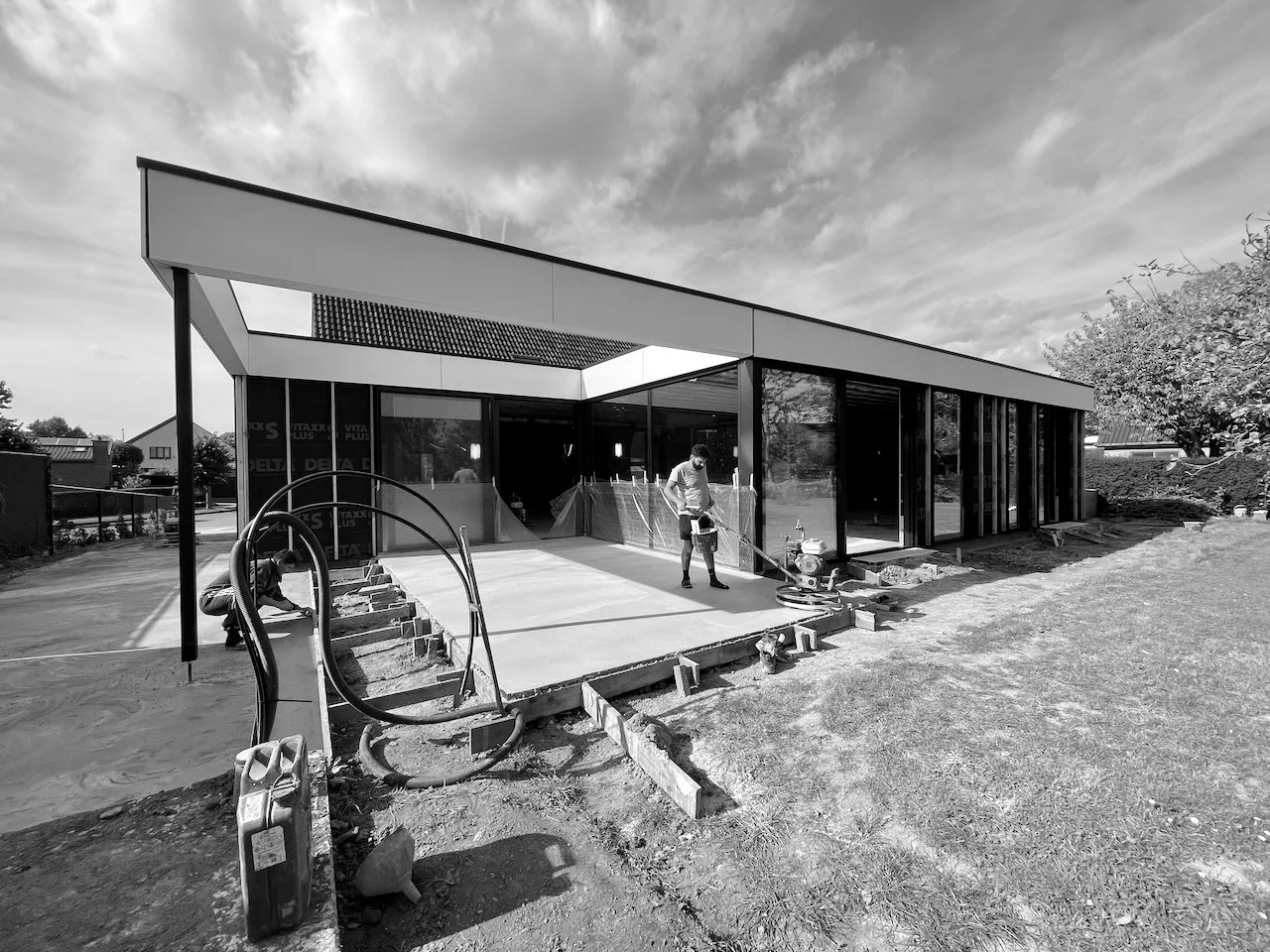 Een gebouw in aanbouw met twee bouwvakkers die de vloer afwerken, omringd door bouwmaterialen en gereedschap, onder een bewolkte hemel.