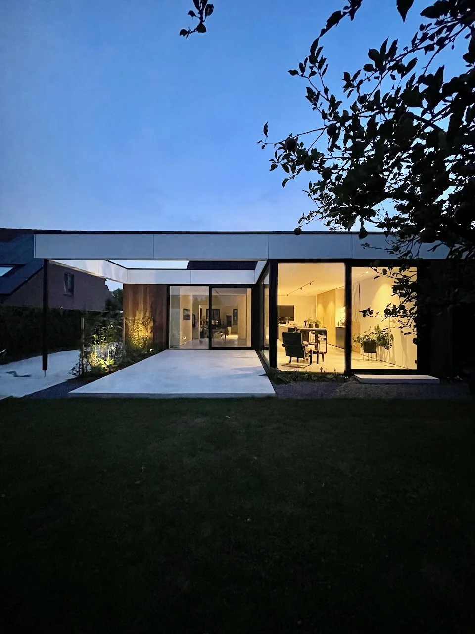Moderne huis met grote glazen ramen en een verlichte interieur, gezien tijdens de schemering, met een grasveld voor en een boom aan de rechterkant.
