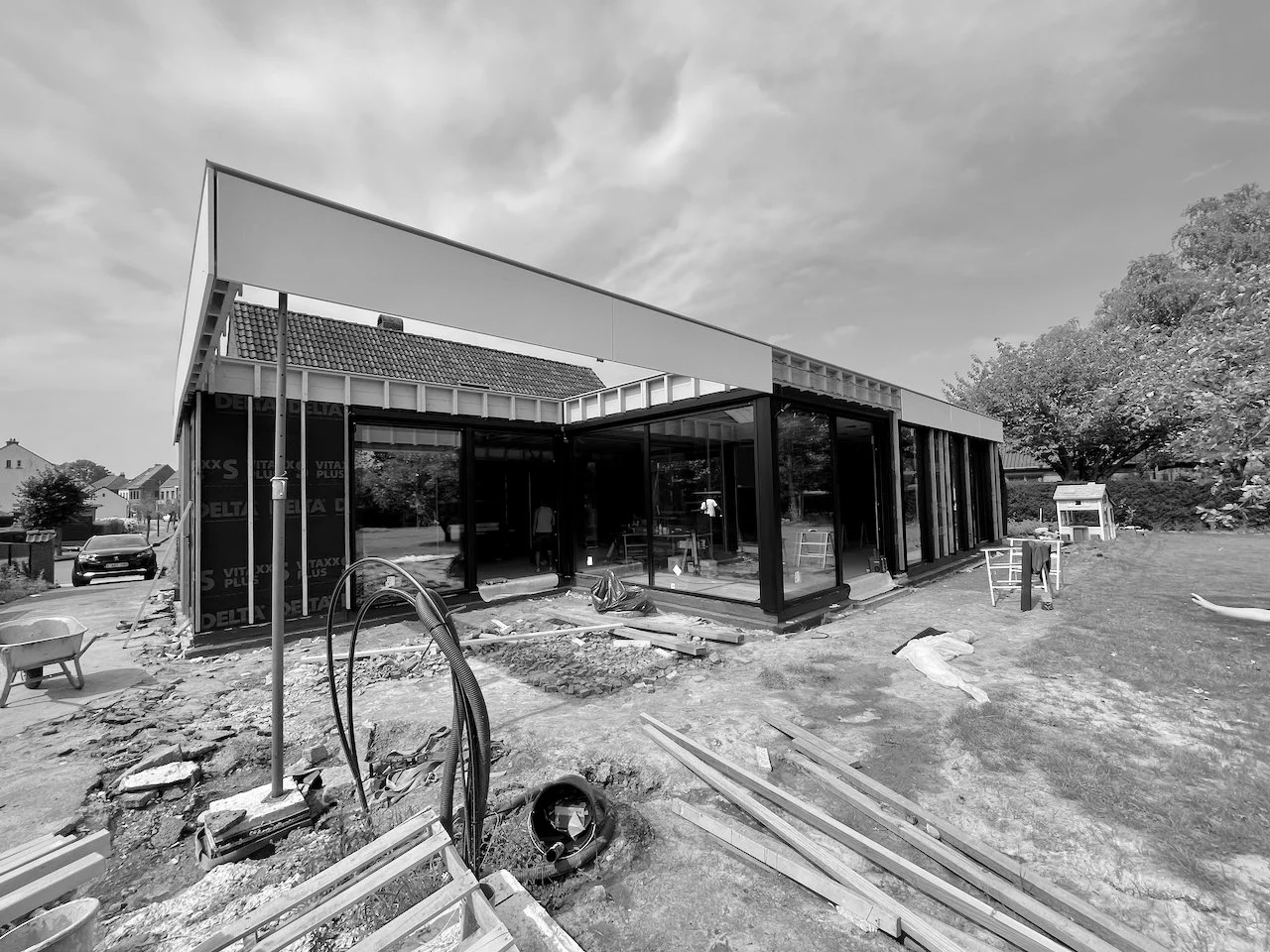 Een moderne bouw van een gebouw met grote glazen ramen, omgeven door bouwmaterialen en gereedschap, met een tuin en bomen op de achtergrond.