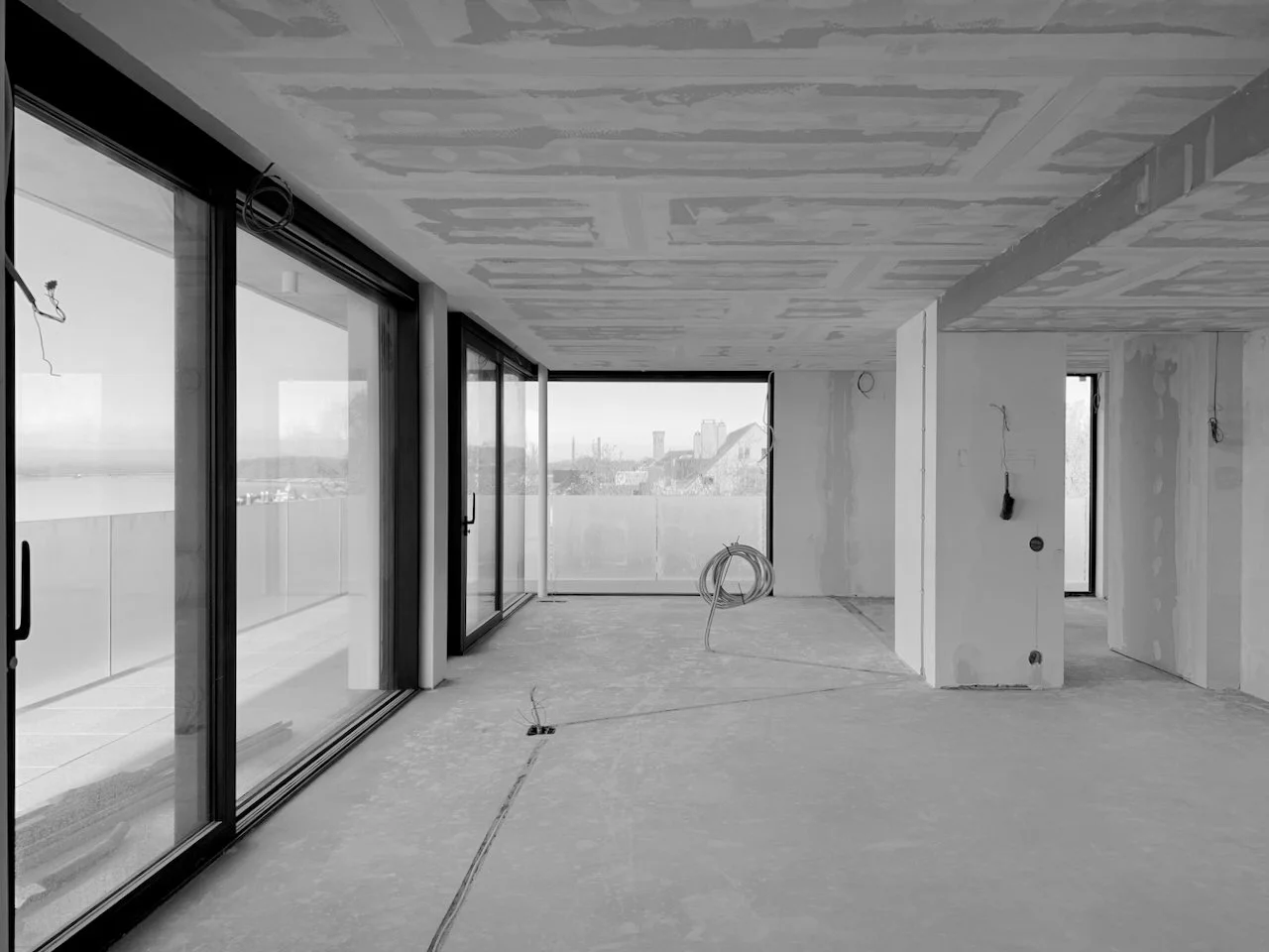 Een lege, onafgewerkte kamer in een hoogbouwgebouw met grote glazen schuifdeuren en uitzicht op een stad.