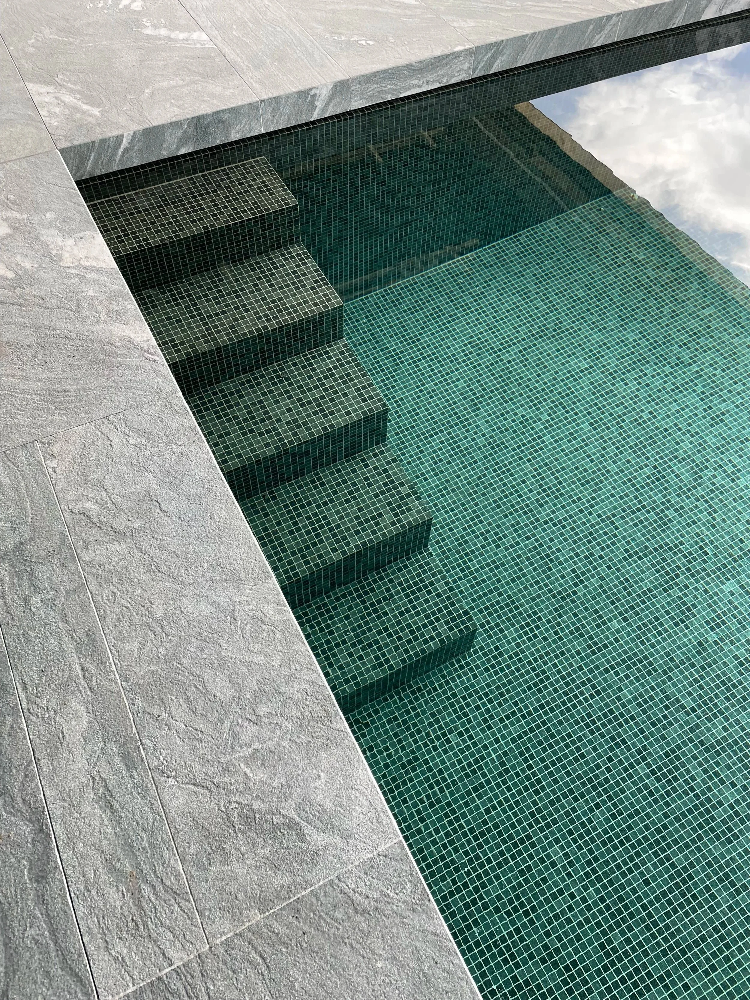 Een zwembad met blauwe mozaïektegels en stenen omlijsting, gezien van bovenaf.