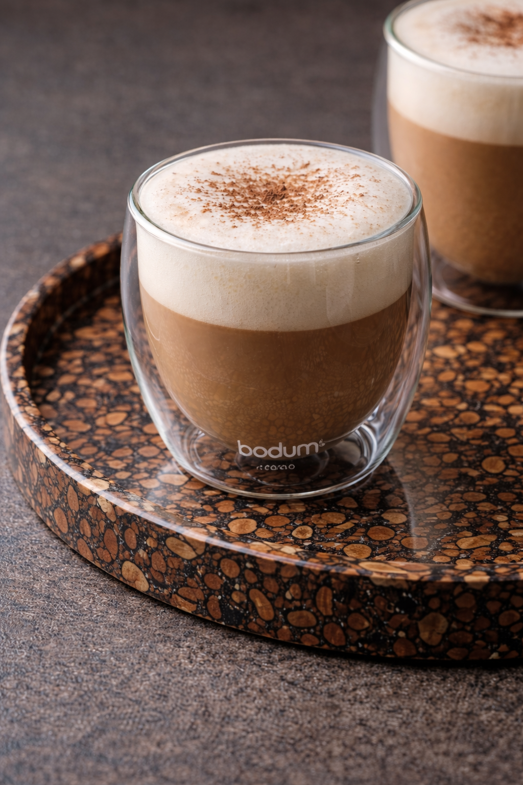 Twee glazen koffie met schuim en cacaopoeder, op een ronde gevlekte houten schaal