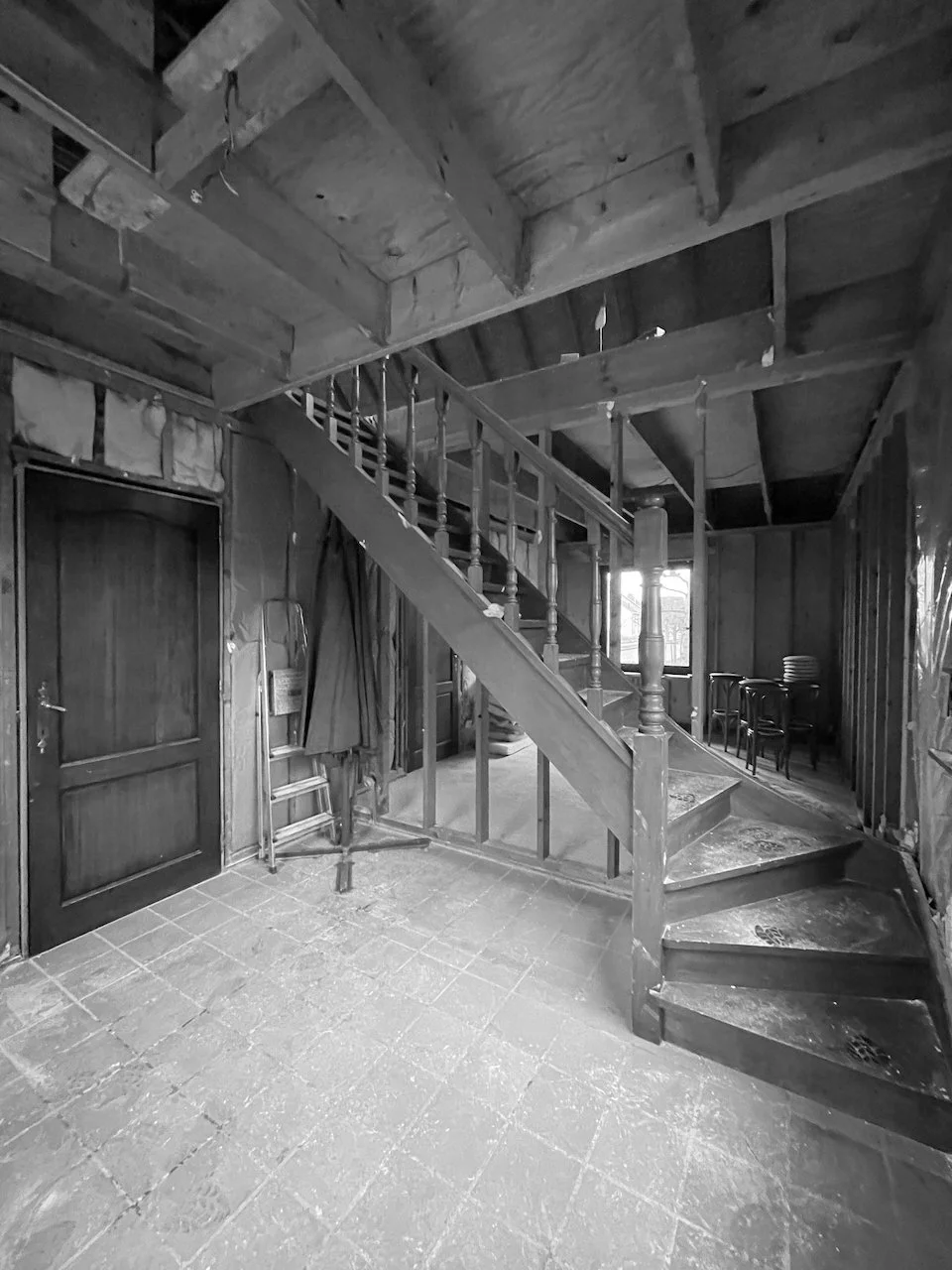 een binnenruimte met een houten trap en houten plafond, een deur links, stoelen in de hoek, en een vloer van tegels, in zwart-wit