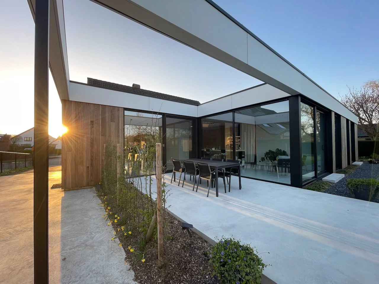 Moderne woning met grote glazen ramen en patio, met een tuin en zomerse zonsondergang op de achtergrond.
