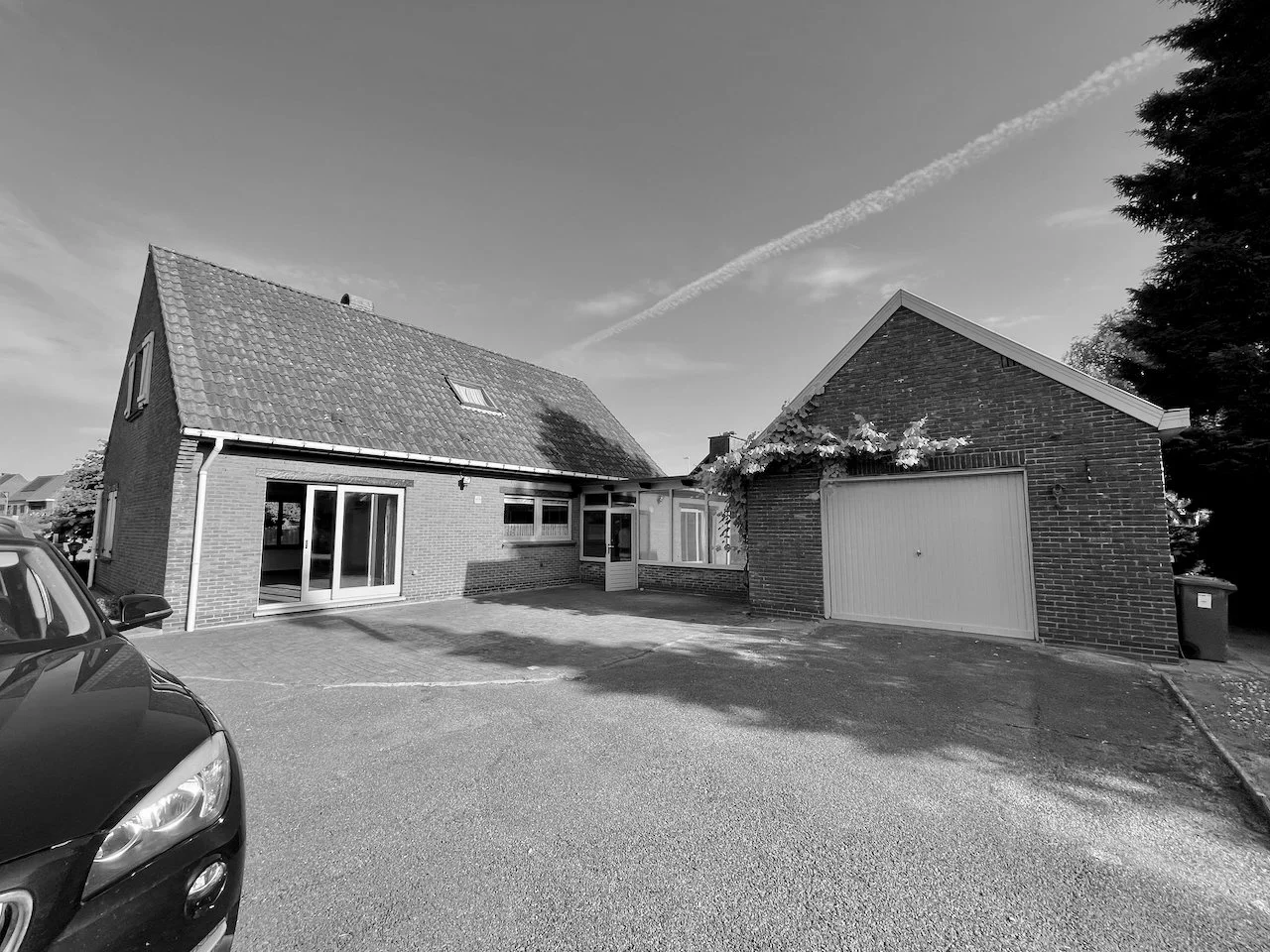 Een zwart-wit foto van een huis met een garage, een auto op de oprit, een grote boom en een heldere hemel met een wolk en een vliegtuigspoor.