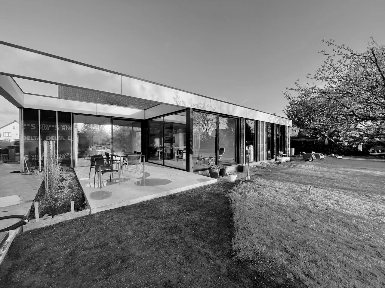 Moderne glazen gebouw met buitenzitje en tuin, mogelijk een café of kantoor, met een boom op de achtergrond.