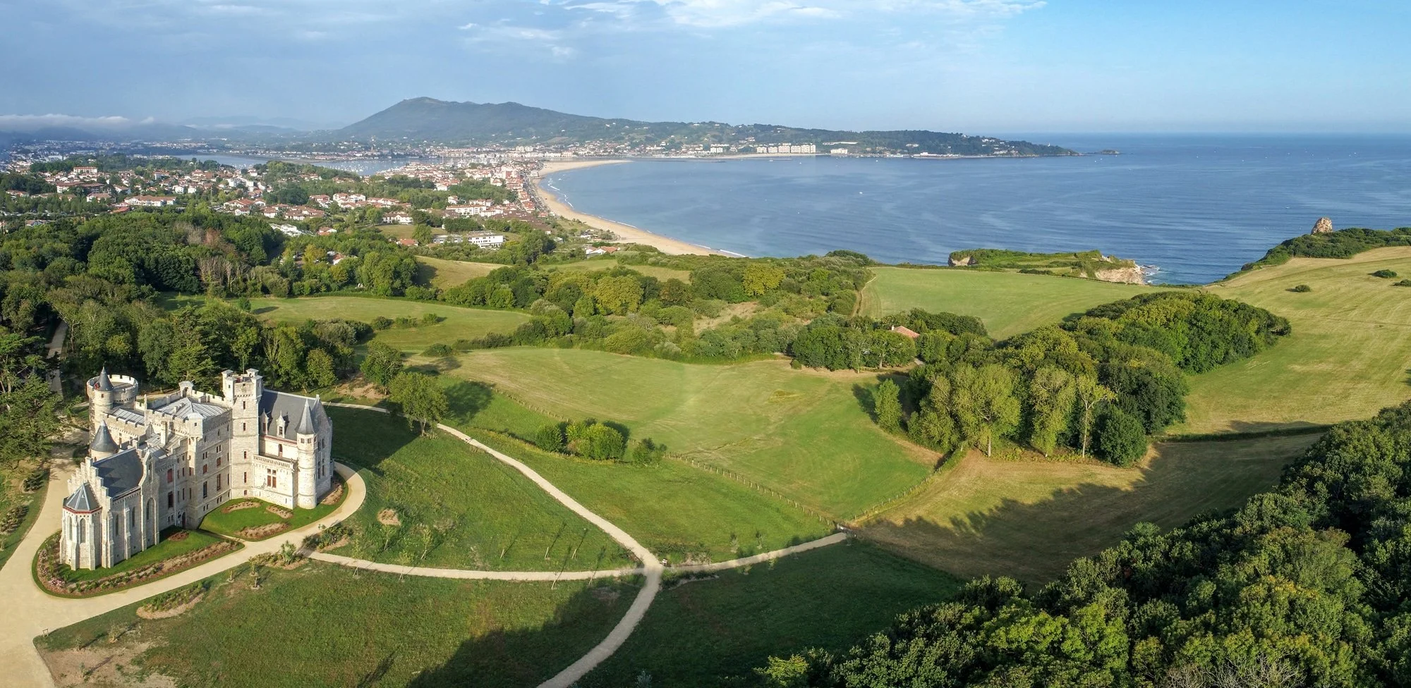 Château blanc situé dans un paysage verdoyant avec des arbres, des champs et une vue sur la mer et une ville en arrière-plan.