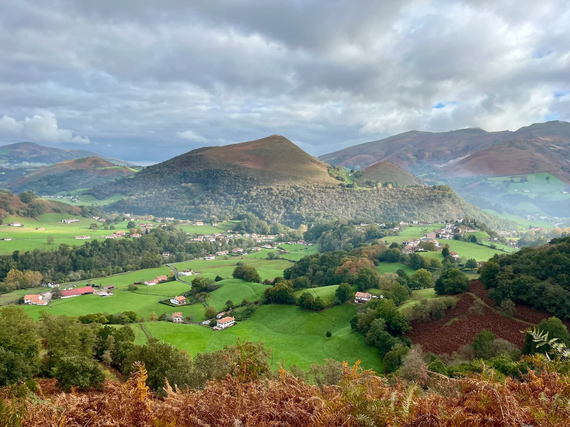 Paysage vallonné avec des maisons éparpillées, des champs verts et des montagnes lointaines sous un ciel nuageux.