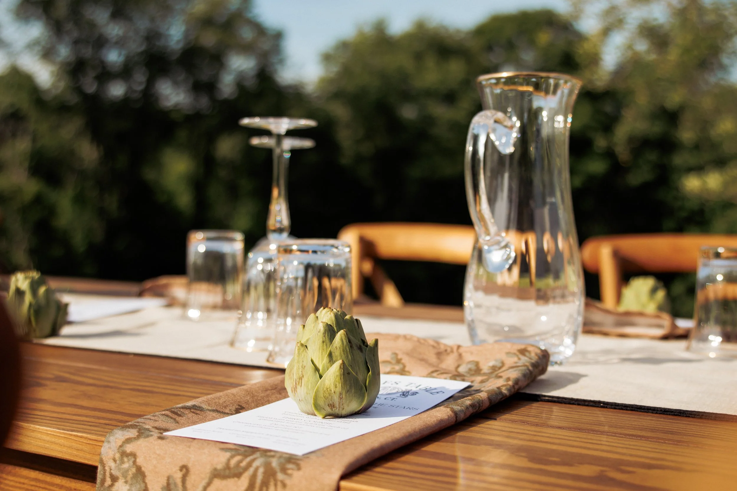 A table set with water pitchers, glassware, artichokes, and a menu on a tablecloth outdoors with trees in the background.