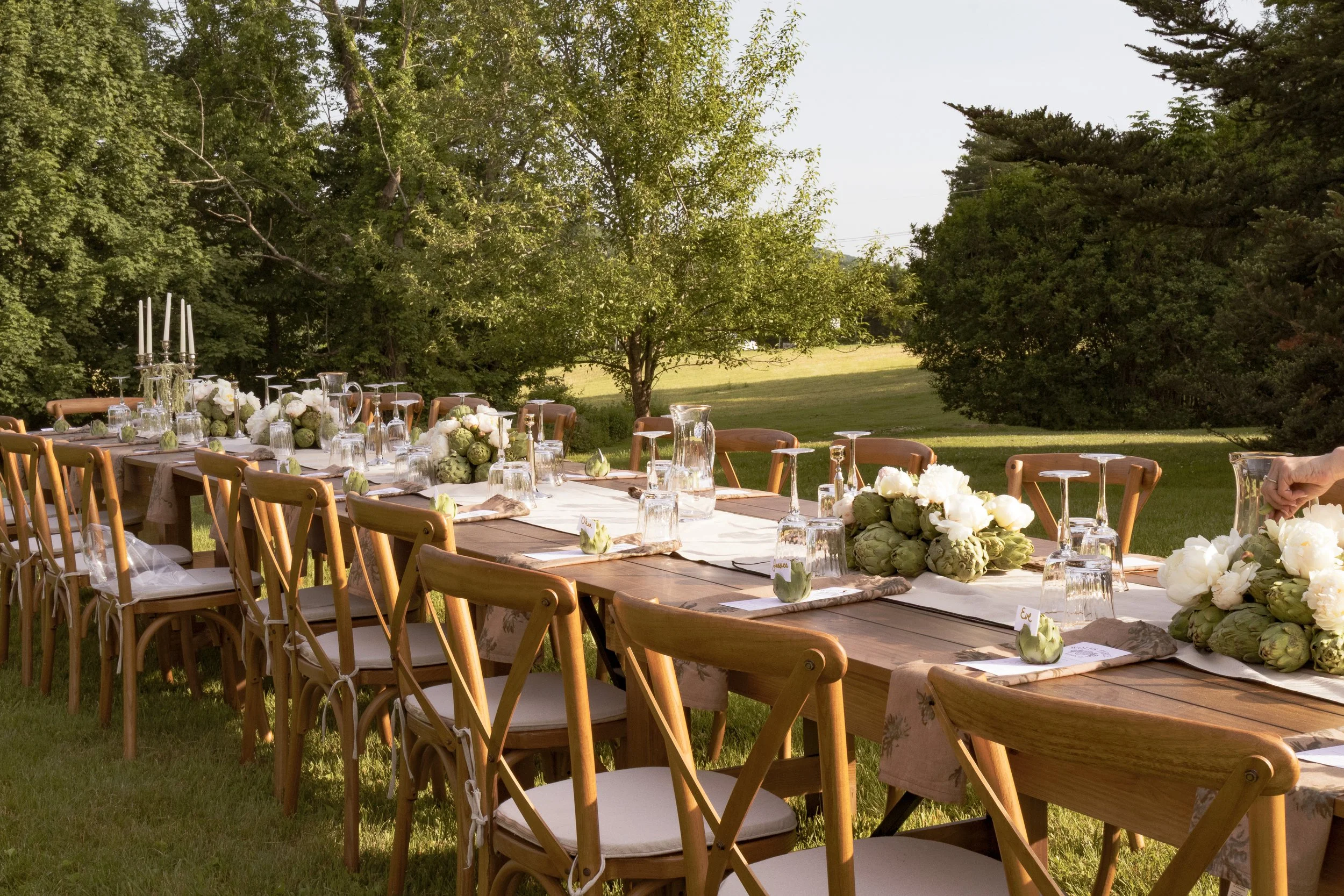 An outdoor dining table set on grass with floral centerpieces and glassware, surrounded by trees under clear skies.