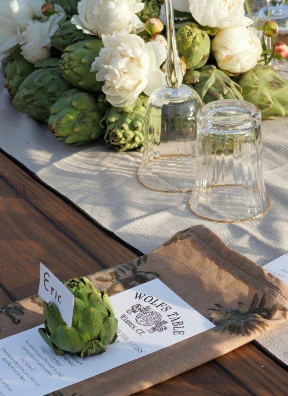 A table setting with white flowers, two upside-down clear glasses, an artichoke with a name card labeled 'Eric,' and a menu from Wolfs Table on a wooden table. The background includes greenery and a white tablecloth.