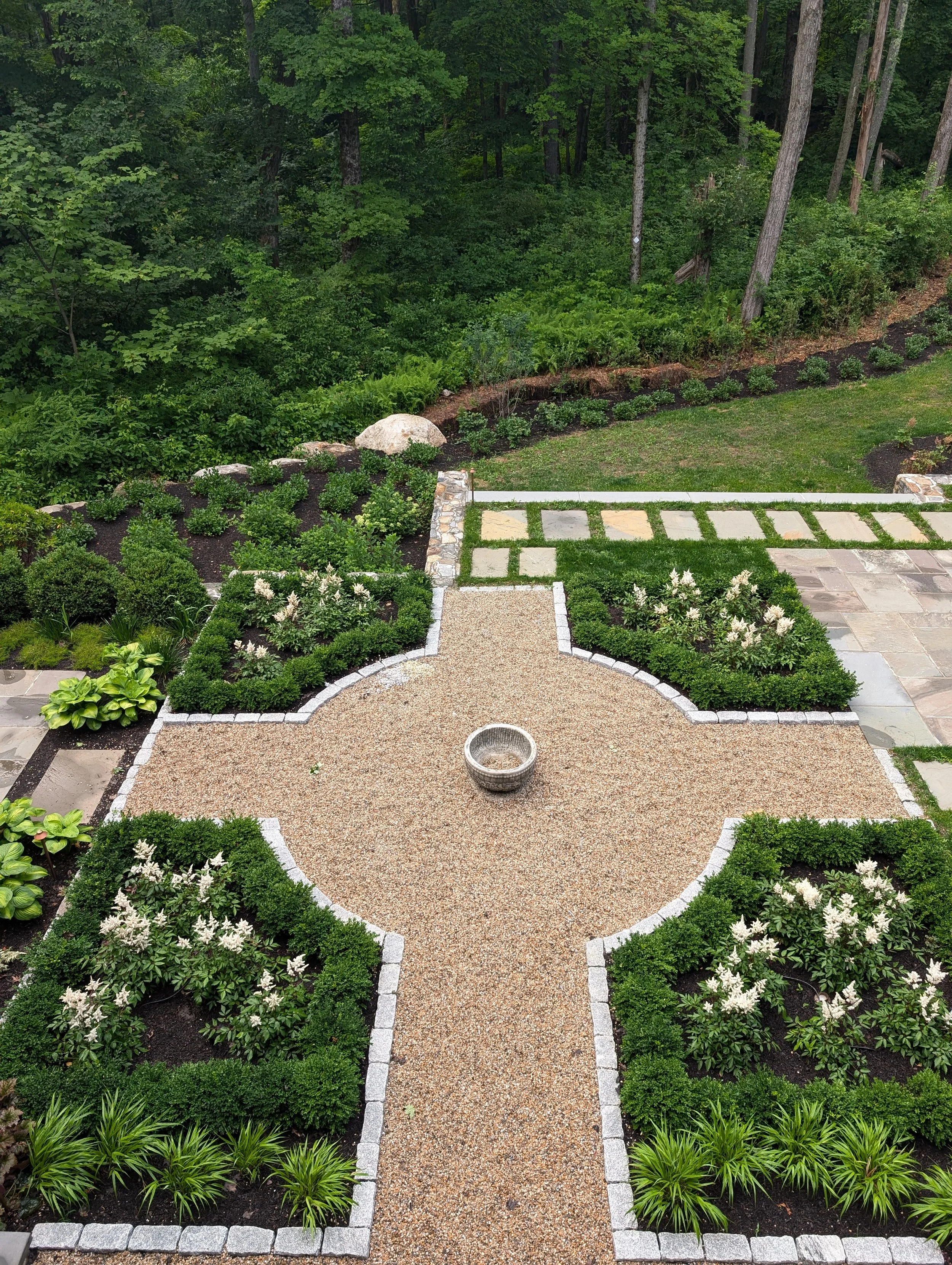 A well-maintained garden with a gravel pathway leading towards a lush green forest. The garden features neatly trimmed bushes and flowering plants, bordered by stone edging, with some large rocks and a decorative stone birdbath in the center.