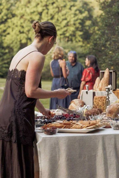 A woman in a black dress serving herself food at an outdoor buffet table with bread and pastries, while three people in the background are talking.