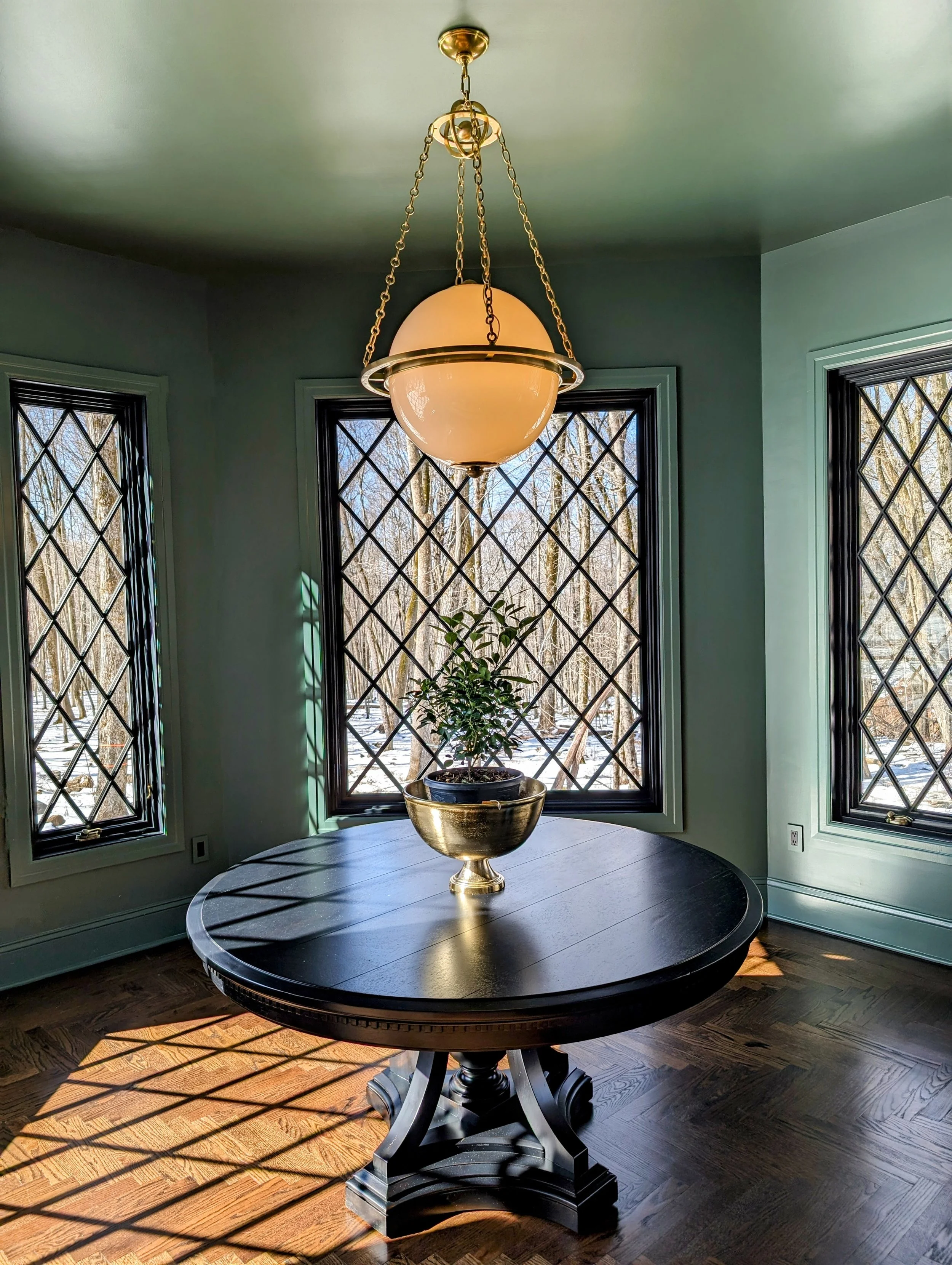 A round wooden dining table with a black finish, featuring a gold bowl containing a potted plant. Behind the table, there are three large windows with black diamond-patterned wrought iron grilles, revealing a snowy wooded landscape outside. A decorative pendant light hangs above the table.
