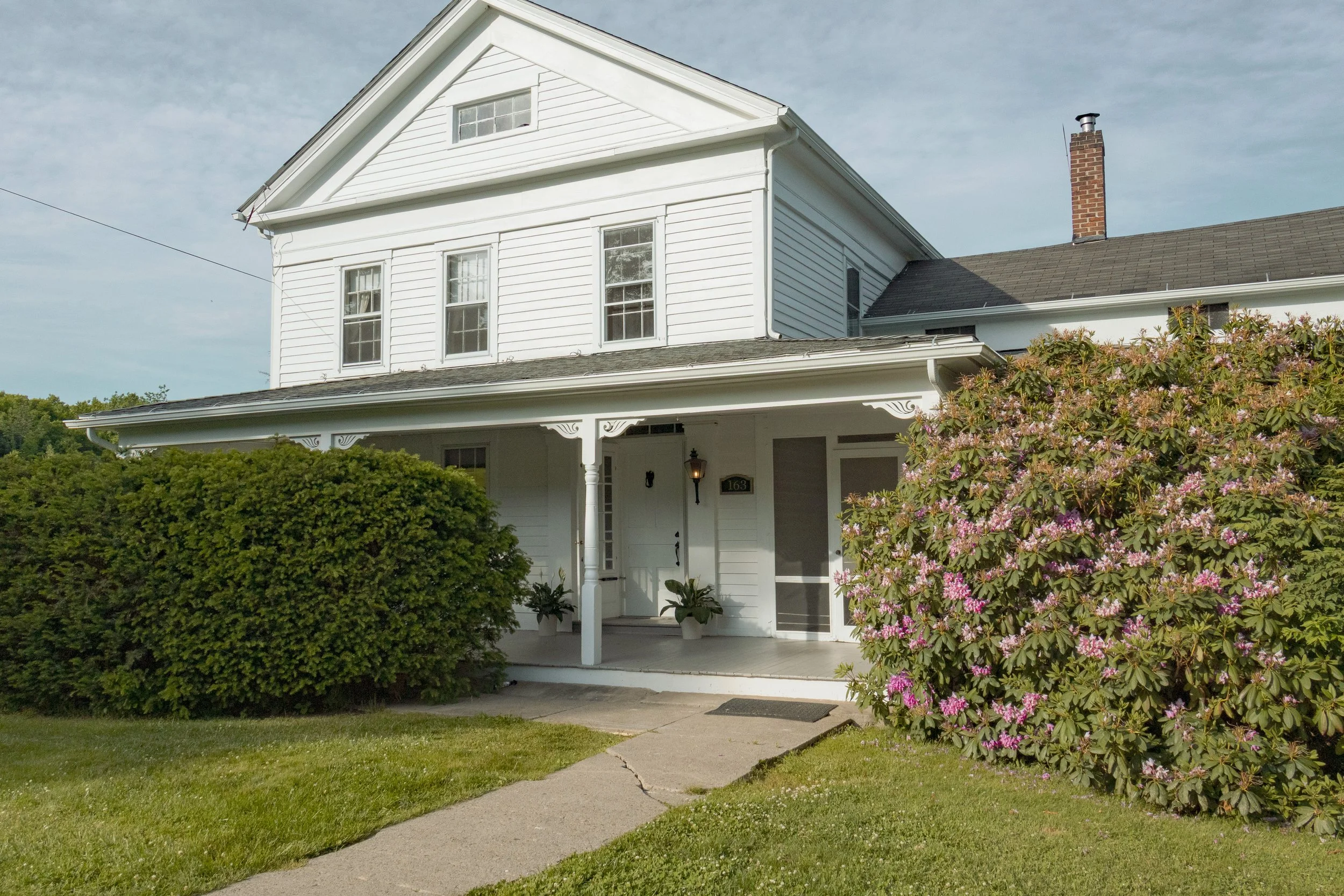 White two-story house with a porch, surrounded by green bushes and pink flowering bushes, under a cloudy sky.