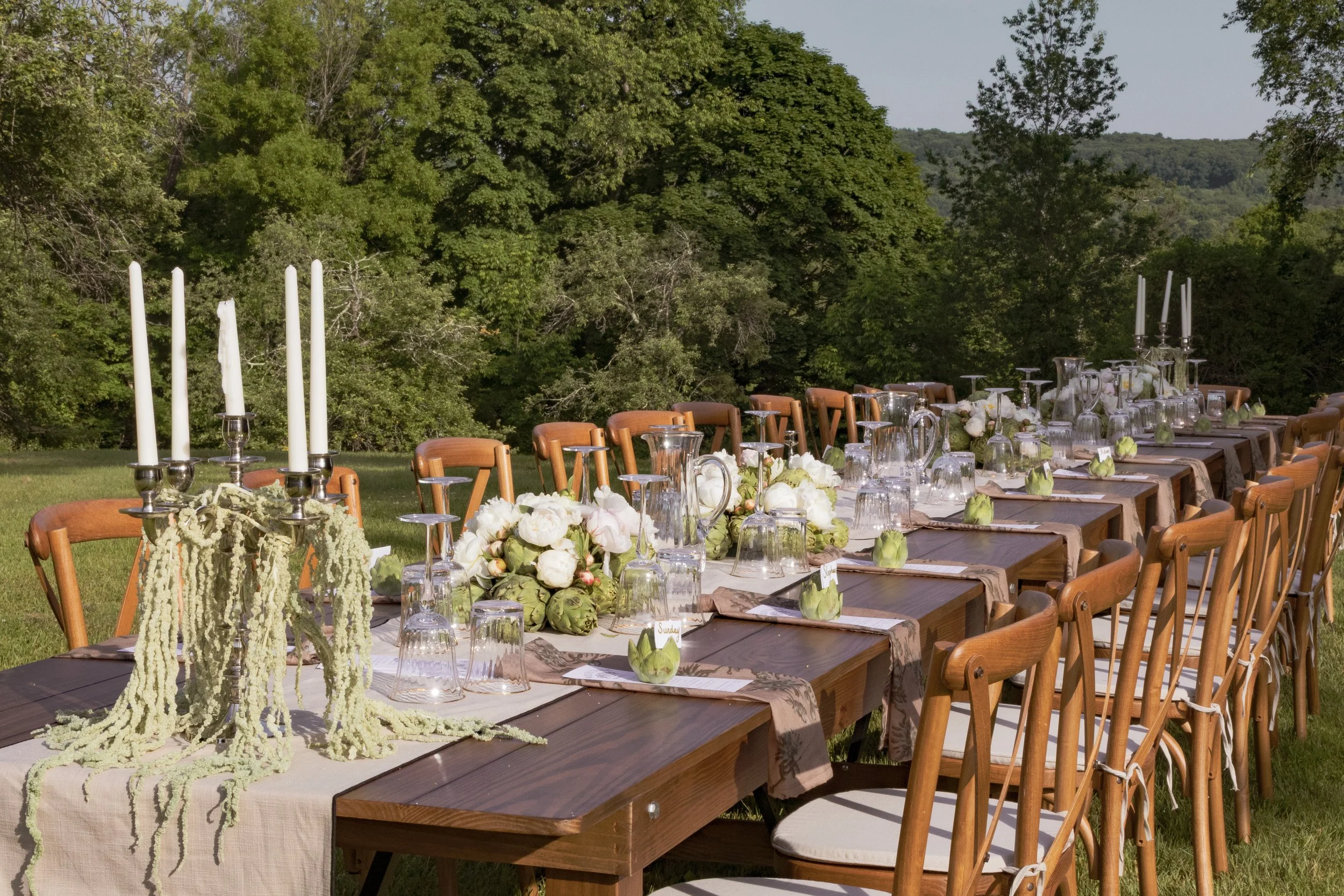 A long dining table outdoors with wooden chairs, white floral centerpieces, glassware, and candles, set for an event in a lush green garden with trees and a mountain in the background.