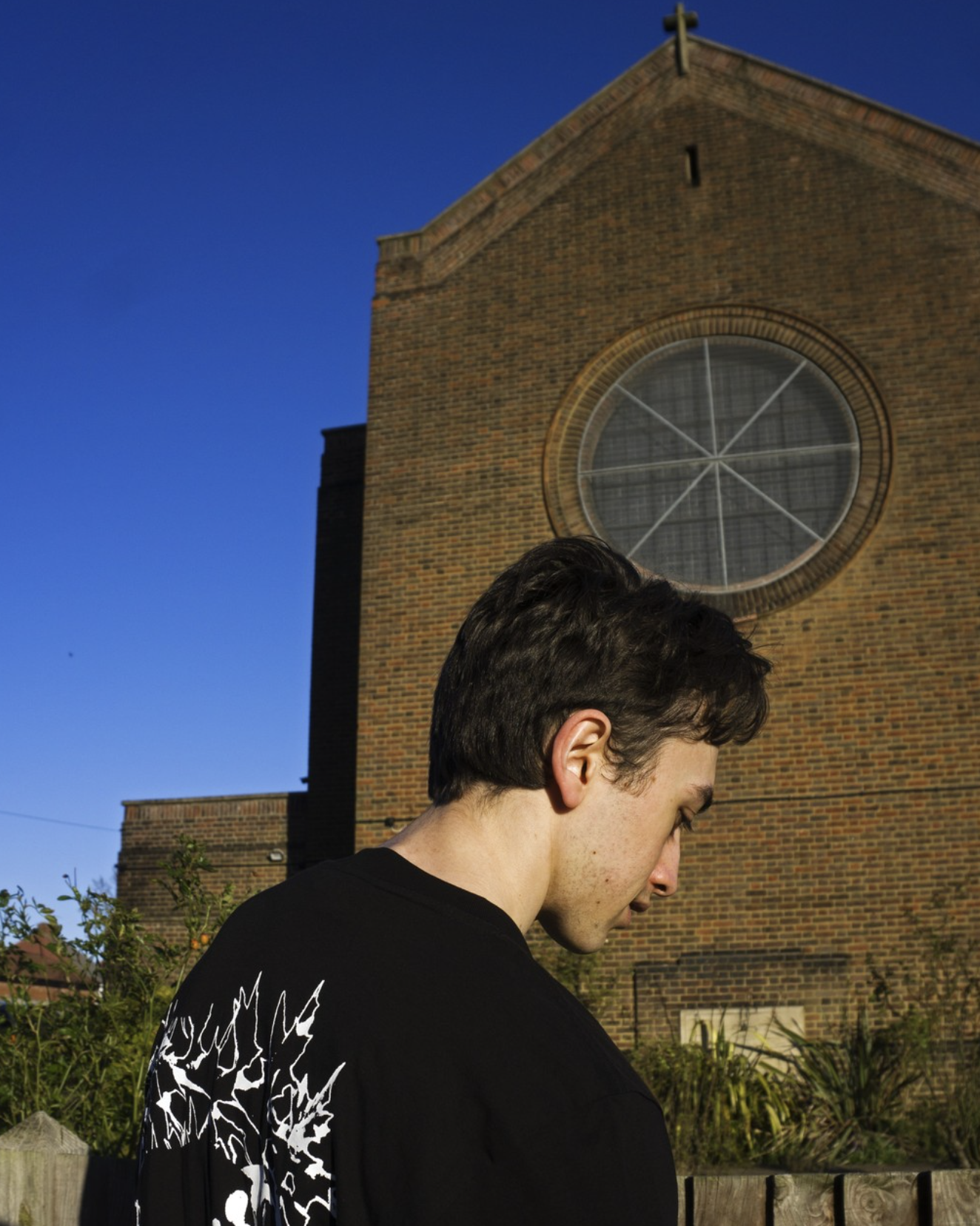 A young man with dark hair looking down outdoors in front of a brick church with a large round window, under a clear blue sky.