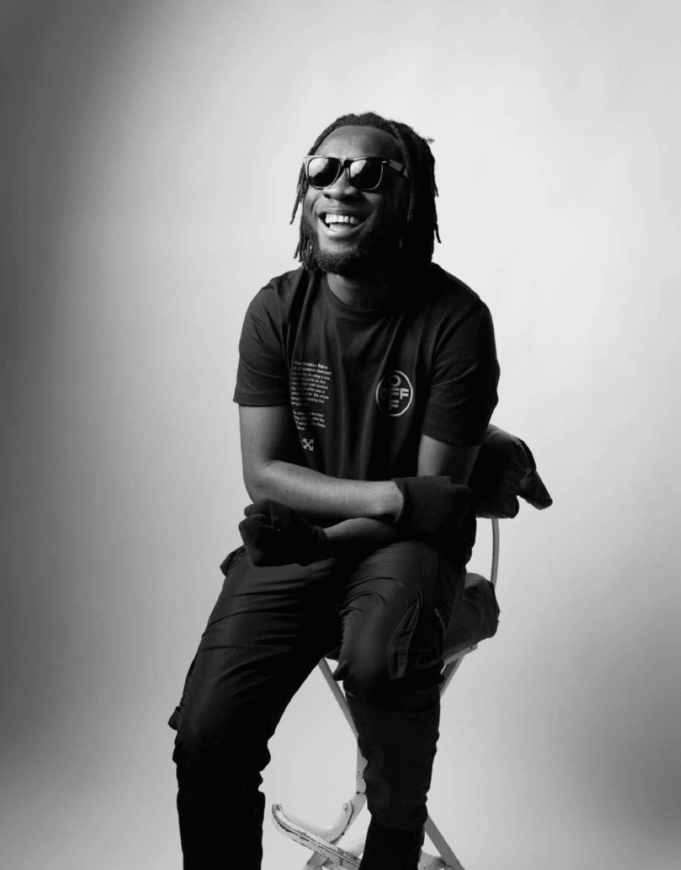 Black and white photo of a smiling man with dreadlocks and sunglasses, sitting on a chair against a plain background.