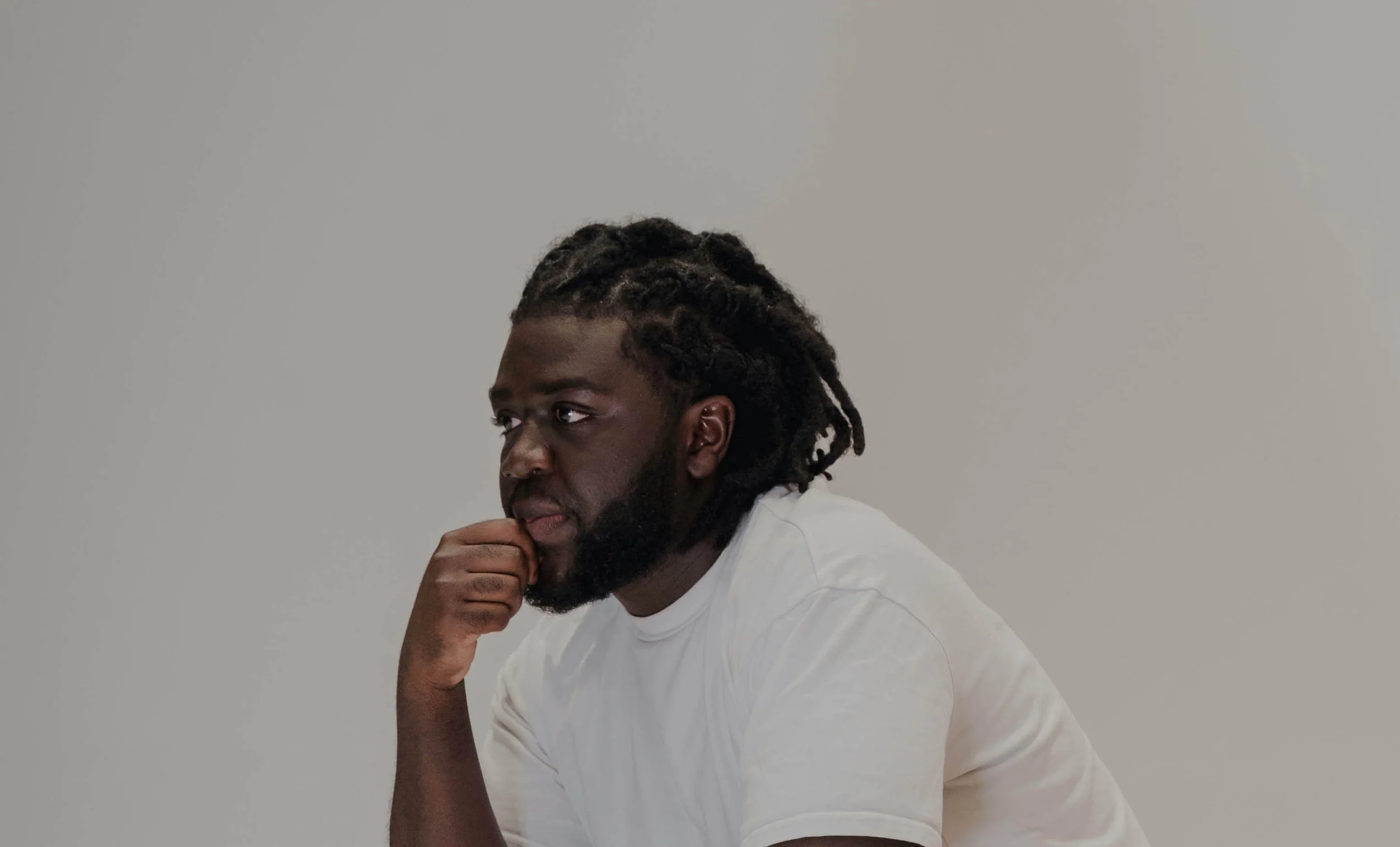 A man with dreadlocks and a beard in a white shirt, resting his chin on his hand, looking to the side against a plain light-colored background.