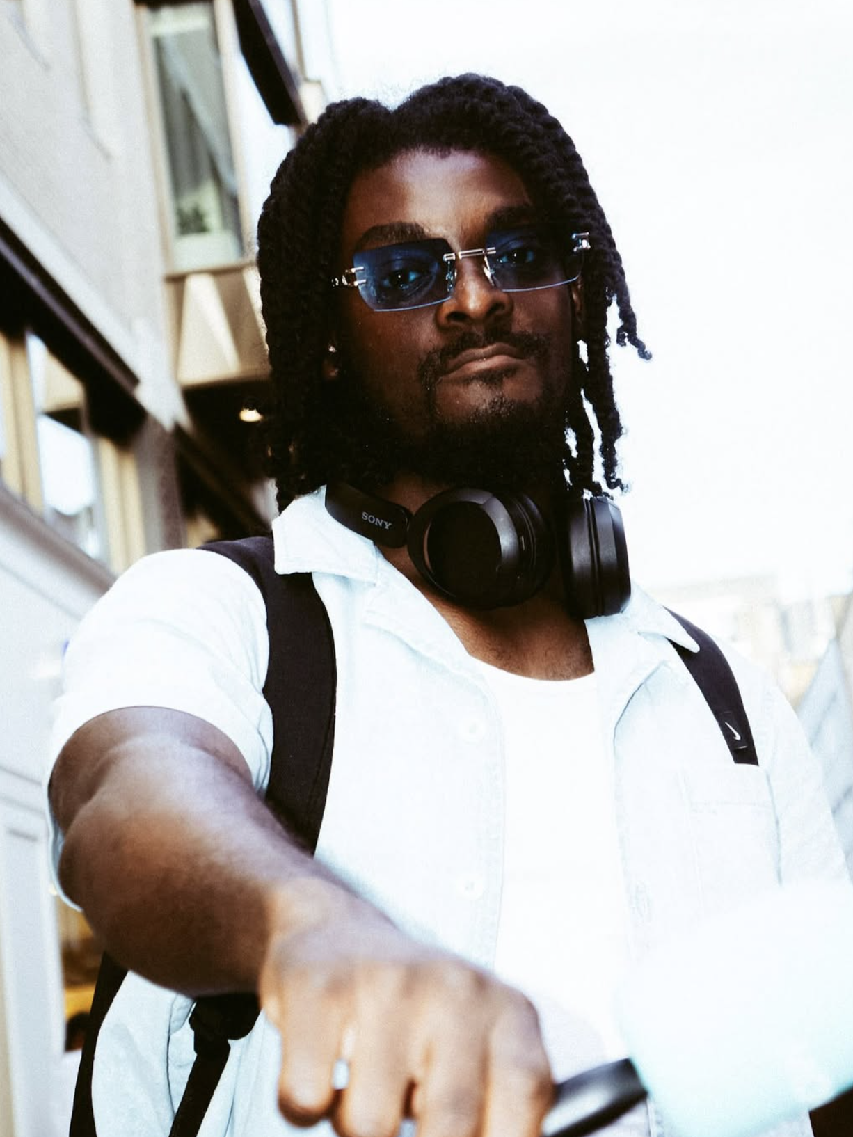 A man with dreadlocks wearing sunglasses, a white shirt, headphones around his neck, and a backpack, taking a selfie outdoors.
