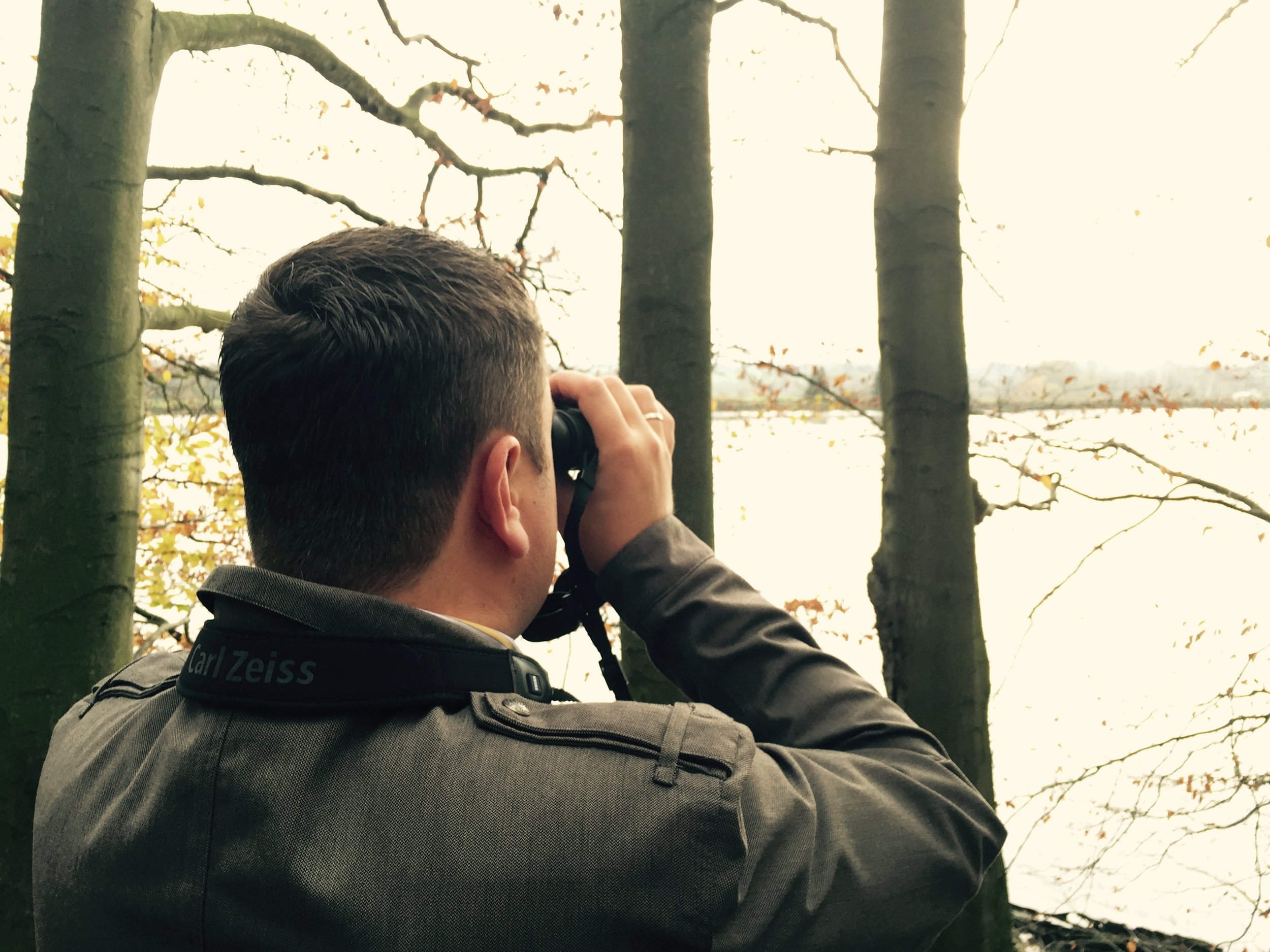 Mann mit Kurzhaarschnitt nimmt ein Foto mit einer Kamera vor einem See auf, umgeben von Bäumen, im Herbst.