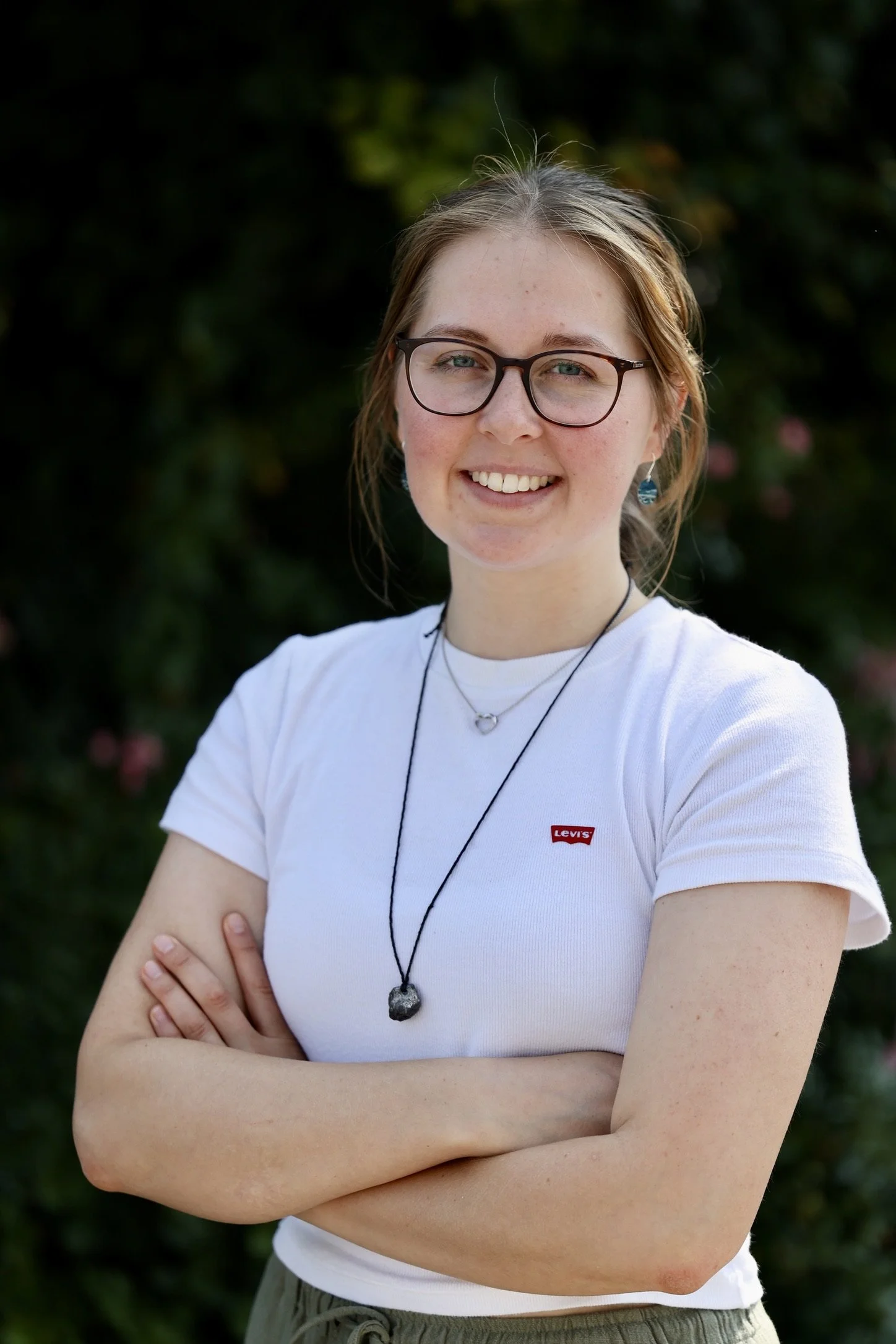 Lächelnde junge Frau mit Brille, braunen Haaren, weißen T-Shirt mit Levi's-Logo, Camping-Sternzeichen-Halskette und Armband, vor grünem Hintergrund.