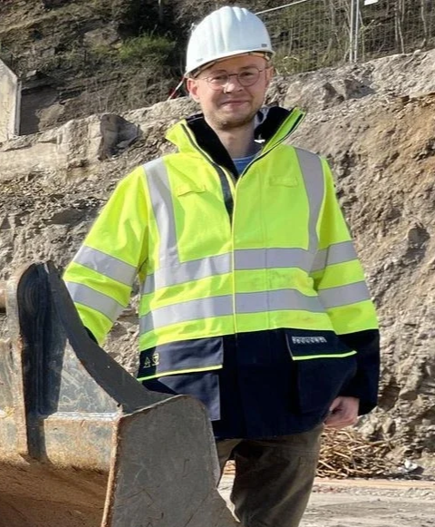 Ein Mann in Sicherheitskleidung und Helm steht vor einem Felsen auf einer Baustelle.
