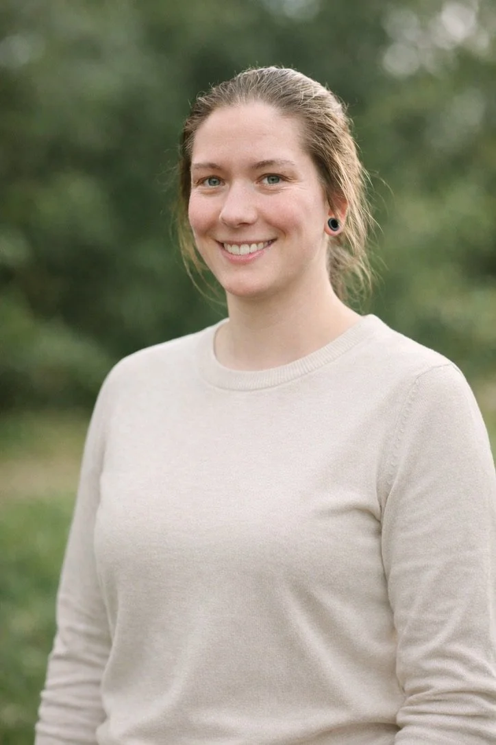 Junge Frau mit braunem Haar und Ohrringen, trägt einen weißen Pullover, steht im Freien vor grüner Natur, lächelt in die Kamera.