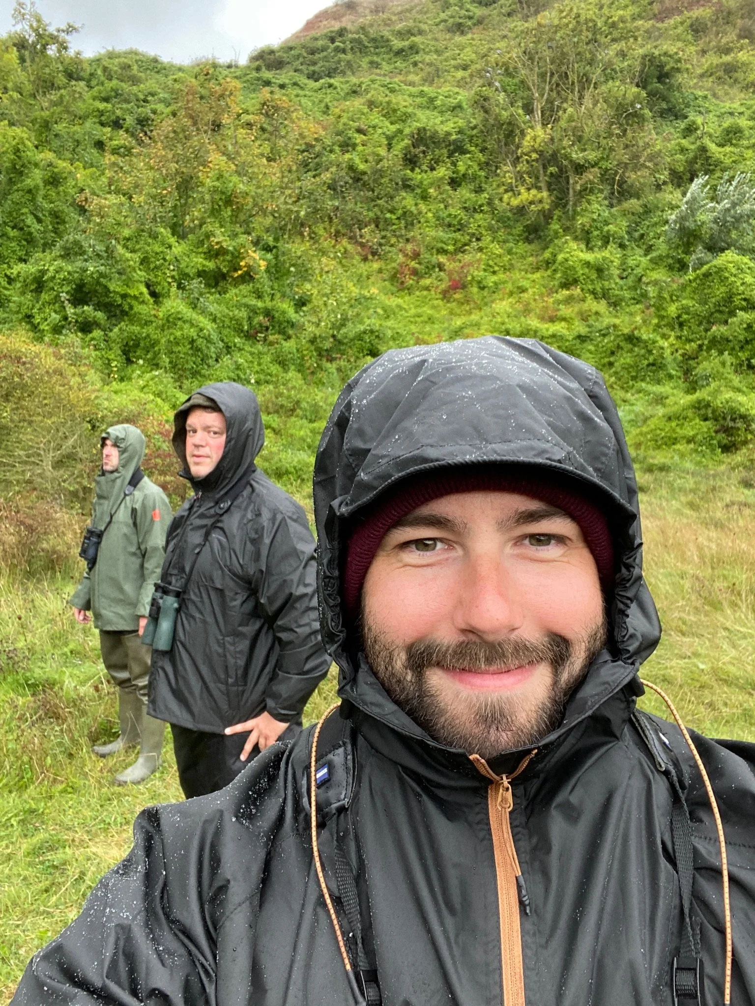 Drei Männer in regnerischer Natur, alle tragen wetterfeste Kleidung mit Kapuzen, der vorderste Mann lächelt in die Kamera.