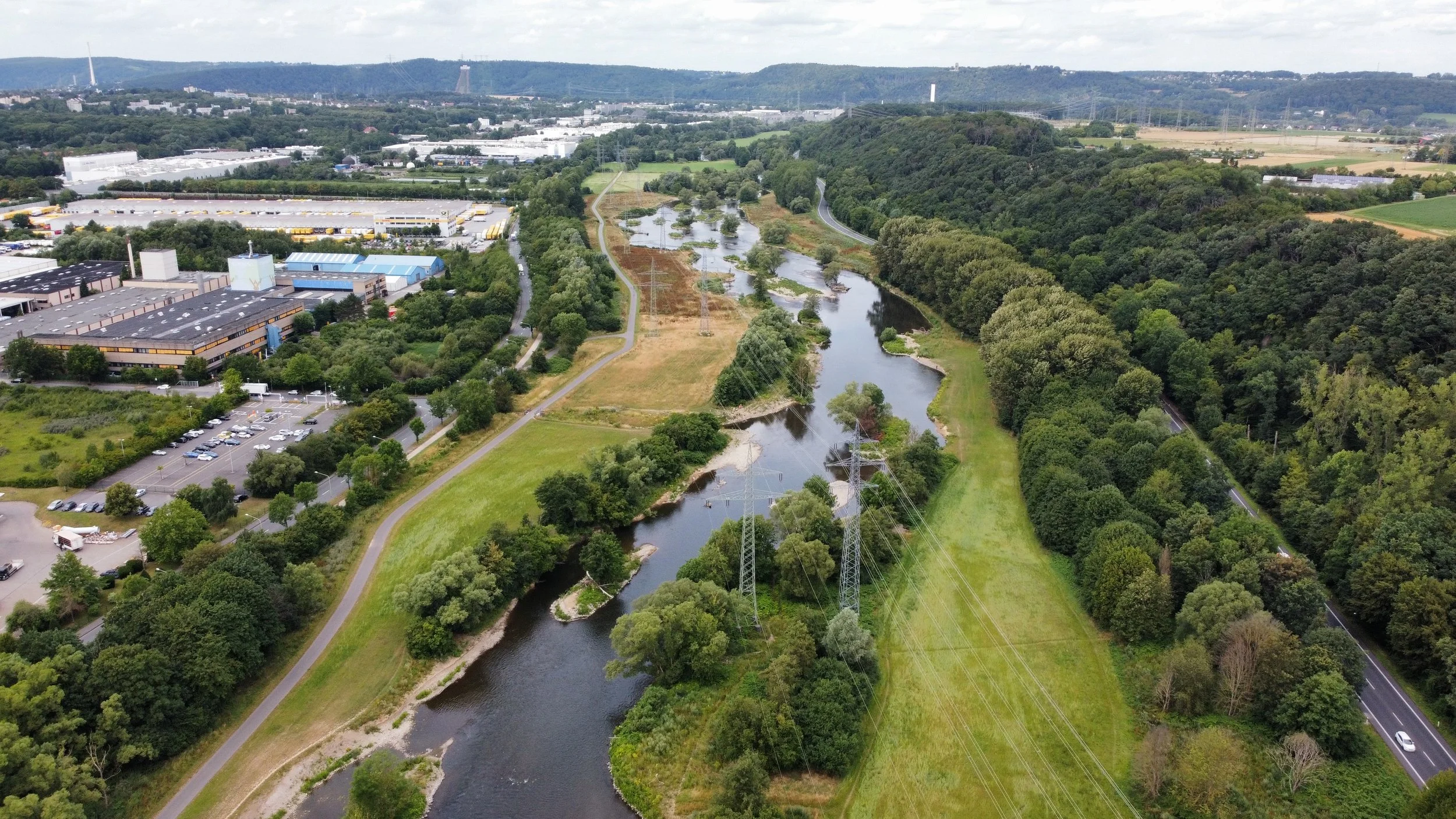 Luftaufnahme eines Flusses, umgeben von Wäldern, Gräsersflächen, Wegen und Straßen, mit Industrie- und Gewerbegebiet im Hintergrund.