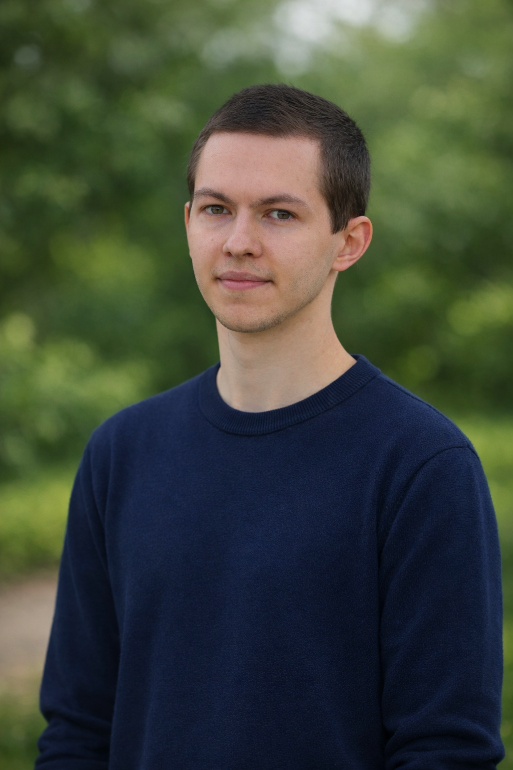 Junge Mann mit kurzen dunkelbraunen Haaren, in dunkelblauem Pullover, steht vor grüner Hintergrund mit Bäumen, Blick leicht zur Seite gerichtet.