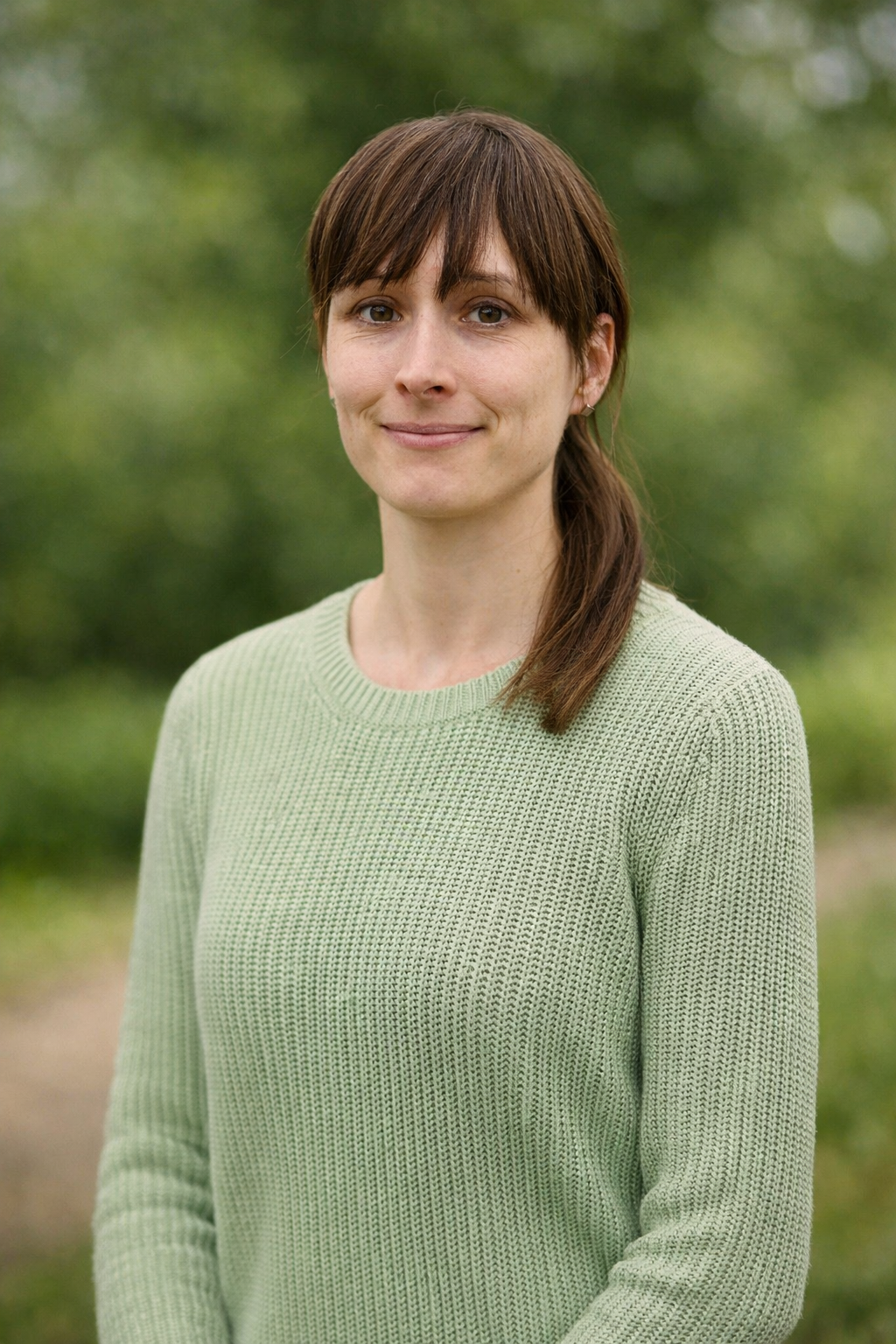 Frau mit braunen Haaren in einem hellgrünen Pullover, lächelnd, vor unscharf grünem Hintergrund im Freien.