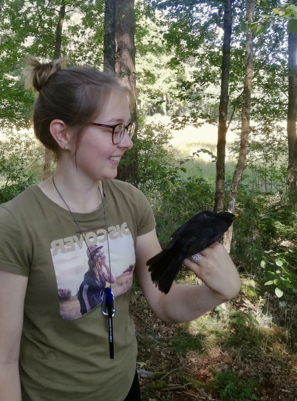 Eine lachende junge Frau mit Brille hält einen schwarzen Vogel auf ihrer Hand in einem Wald, im Hintergrund sind Bäume und grünes Laub.