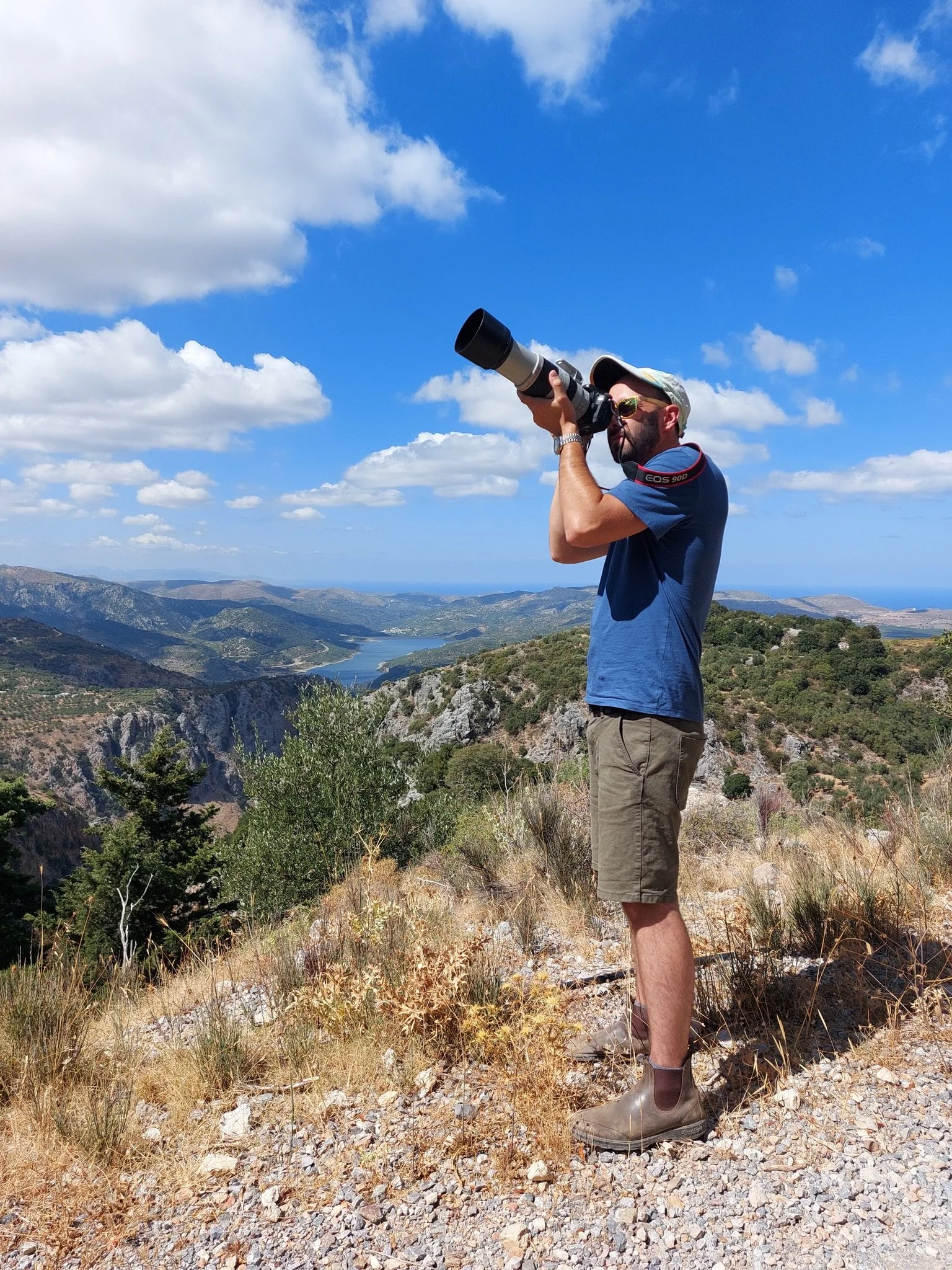 Mann mit Kamera auf einem Hügel mit Berglandschaft im Hintergrund, blauer Himmel und Wolken.