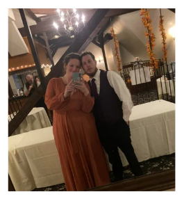 A woman in a long orange dress and a man in a formal outfit taking a selfie in a decorated indoor setting. They are standing in front of a mirror with string lights and orange garlands.