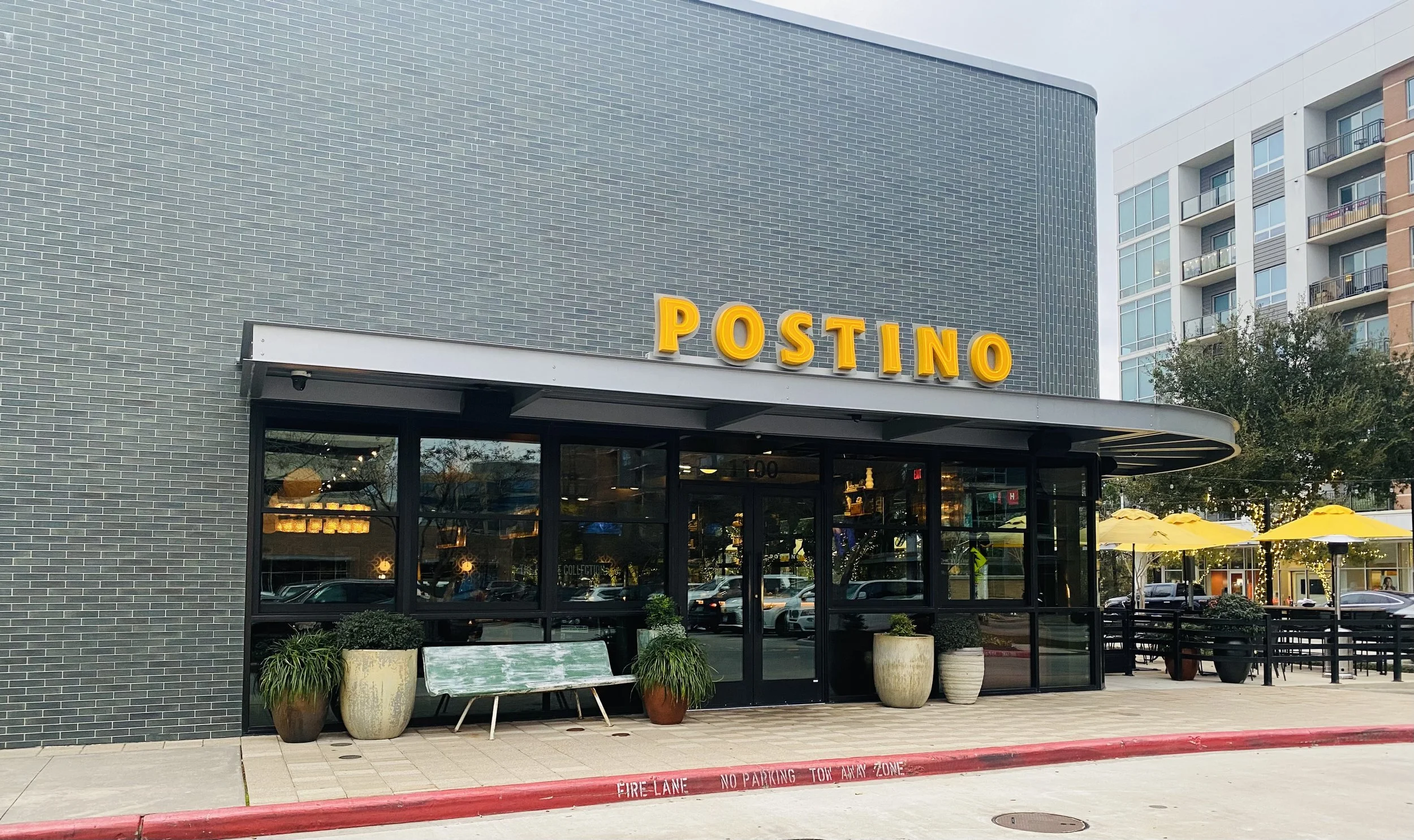 Exterior of a restaurant called Postino with large glass windows, potted plants in front, outdoor seating with yellow umbrellas, and a modern building background.