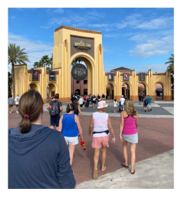 People walking towards the entrance of an amusement park with a large archway and colorful buildings, palm trees, and a blue sky in the background.