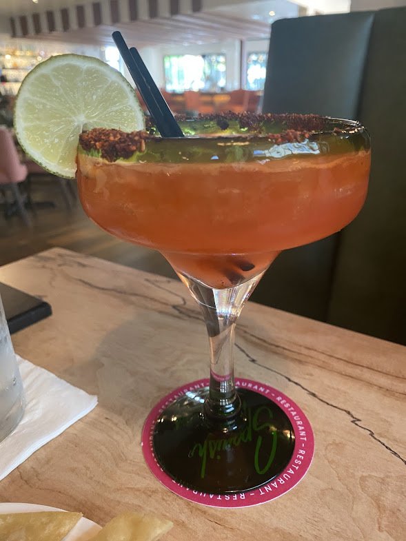 A cocktail in a large margarita glass garnished with a lemon slice and chili powder rim, placed on a restaurant table with a colorful background.