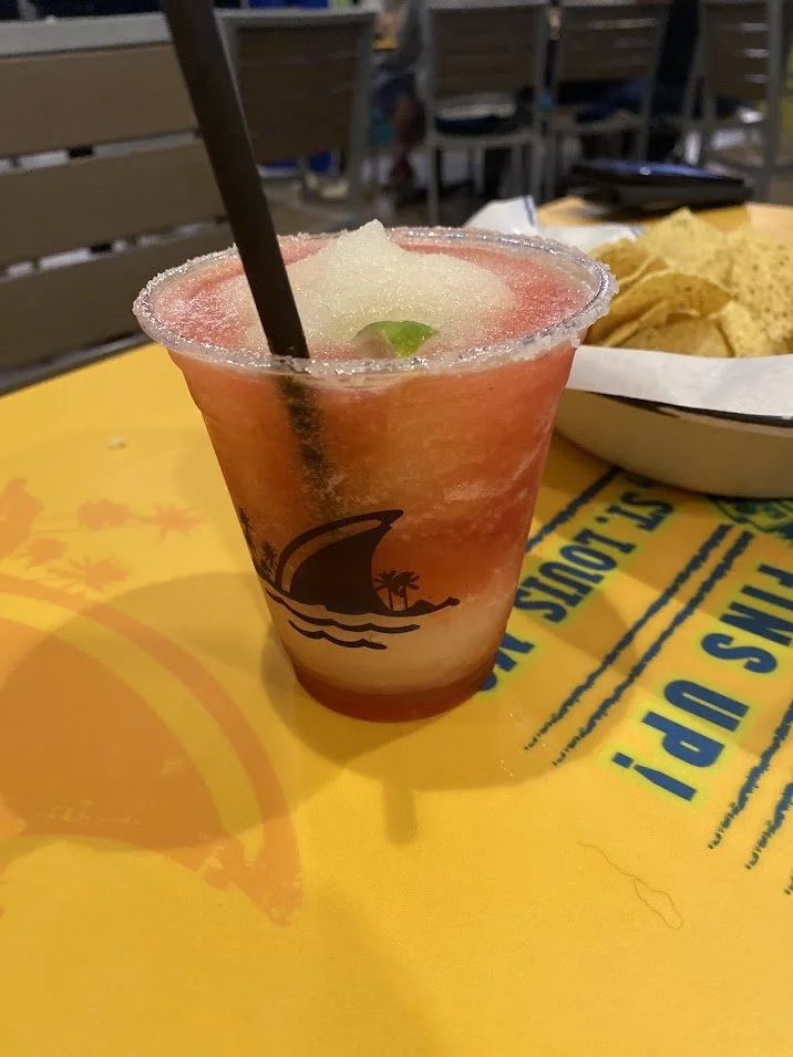 A frozen cocktail with a red and white slushy appearance, garnished with a lime wedge, served in a clear plastic cup with a straw, on a yellow table at a restaurant or bar.