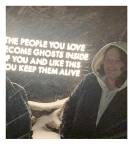 A person smiling, wearing a hoodie, standing near a wall with a quote. The quote says, 'The people you love become ghosts inside of you and like this, you keep them alive.'