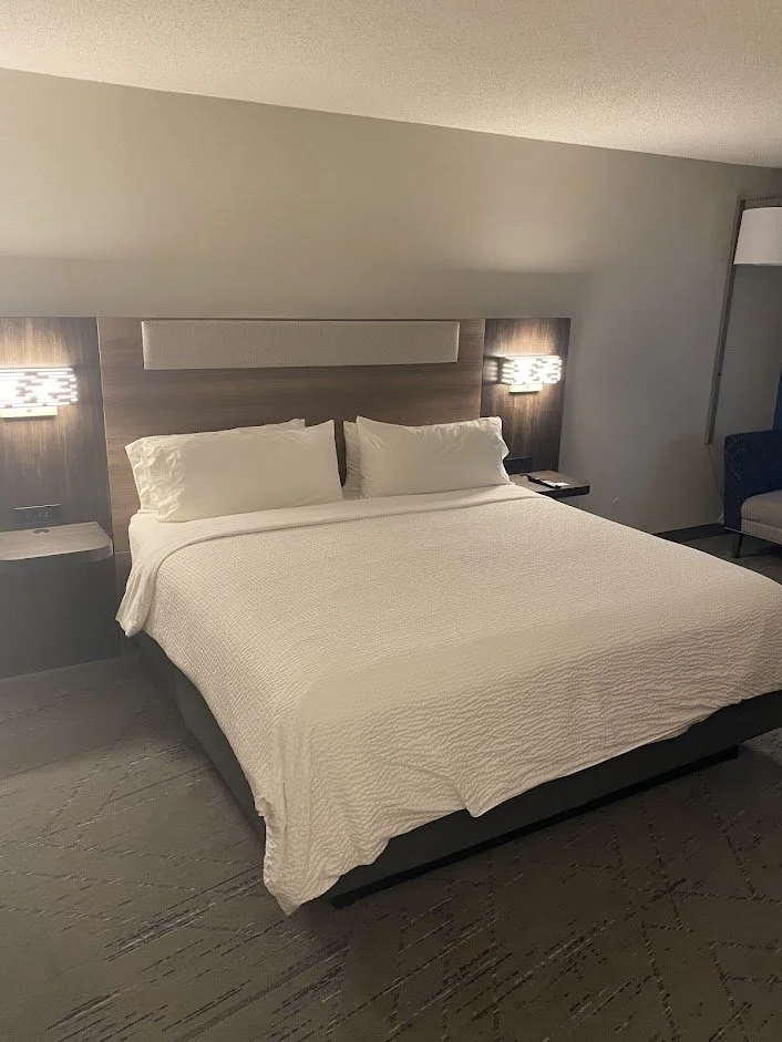 A hotel room with a large bed, white bedding, two pillows, wooden headboard, two wall-mounted lamps, a side table with a phone, and a gray armchair in the corner.