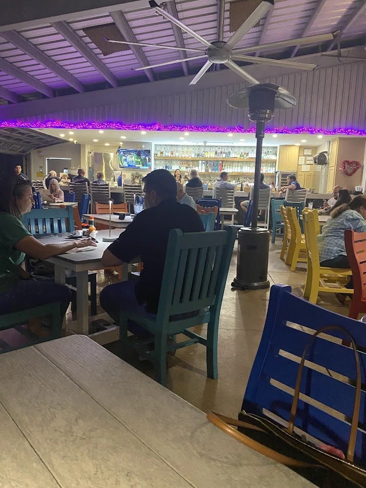 Interior of a lively restaurant or bar with colorful chairs, multiple patrons dining, a bar with bottles, a wall-mounted TV, and a ceiling fan. Purple string lights add ambiance, and a large outdoor heater stands in the middle.